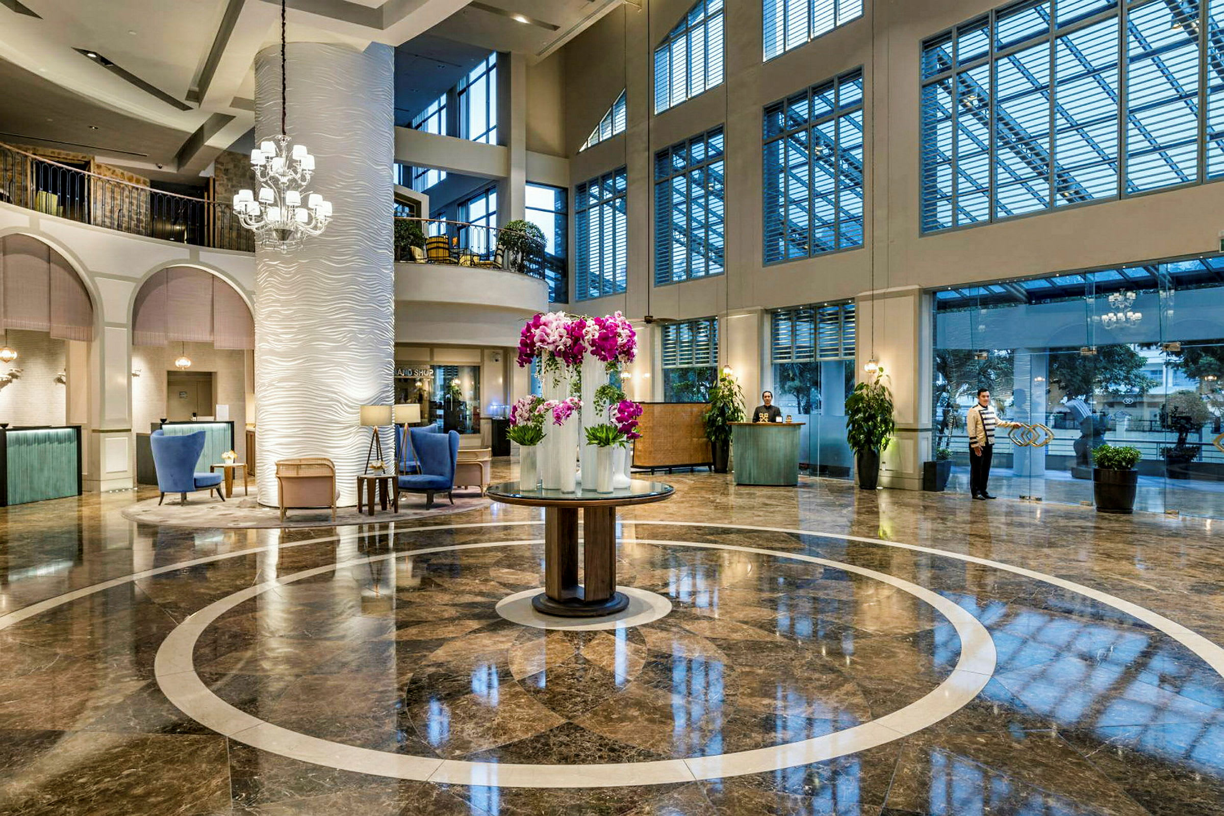 Ett elegant hotellobby med glansig marmorgolv och en central rund plats dekorerad med lila blommor i vaser. Höga fönster släpper in ljus, och en reception syns i bakgrunden. (AI-genererad text)