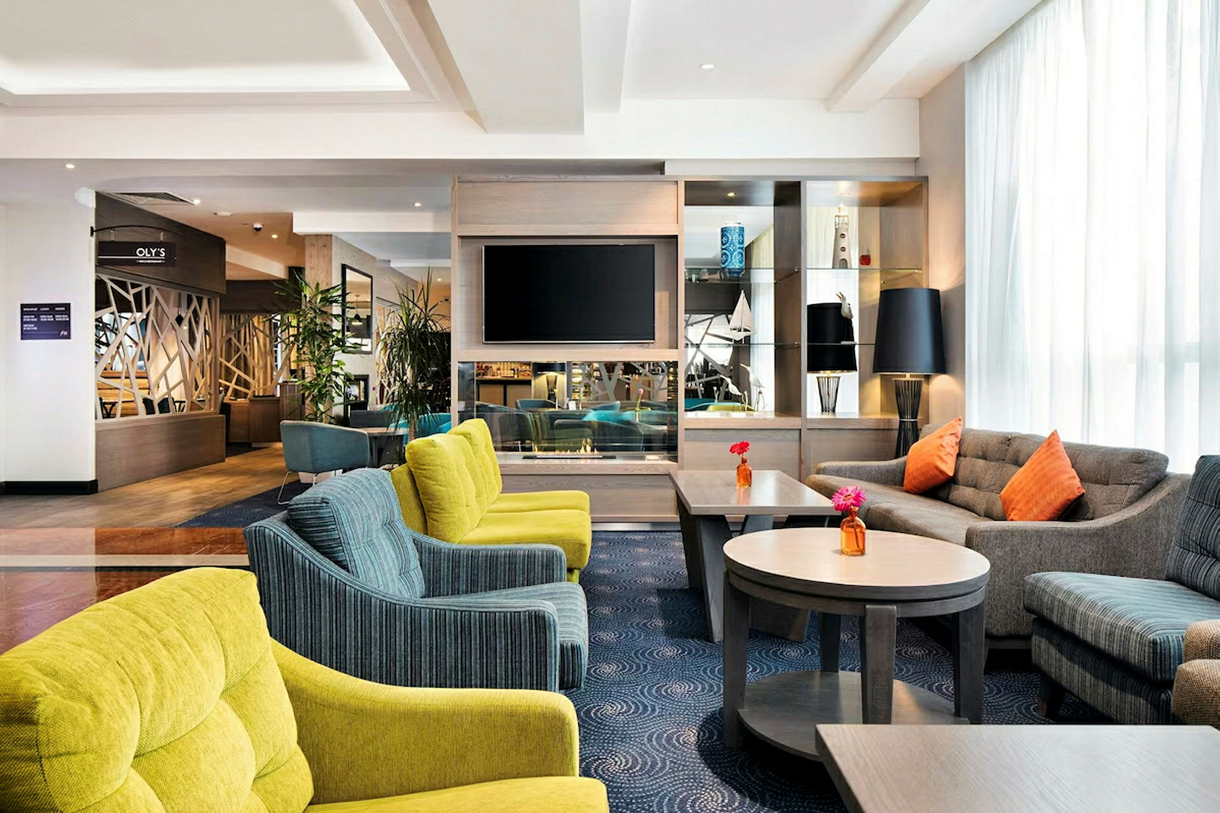 En hotellobby med bekväma sittplatser, flera soffor och stolar i olika färger, samt bord med blommor. En TV är monterad på väggen och det finns dekorativa föremål på hyllorna. (AI-genererad text)
