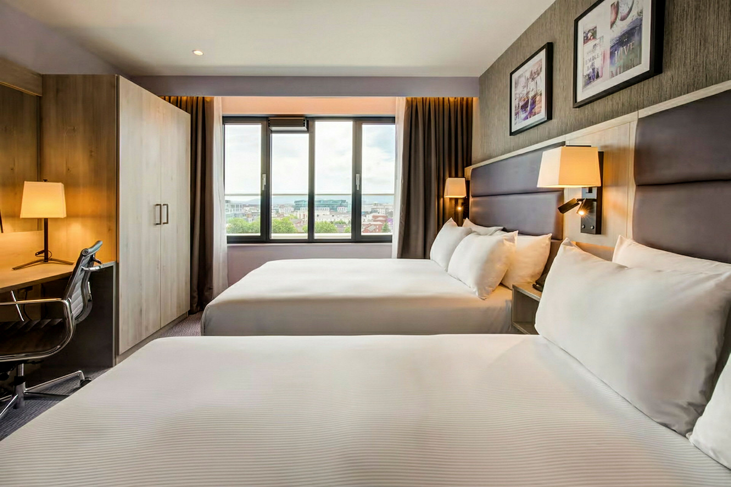 Ett hotellrum med två dubbelsängar, ett skrivbord, en stol, en stor fönsteryta och väggdekorationer. (AI-genererad text)