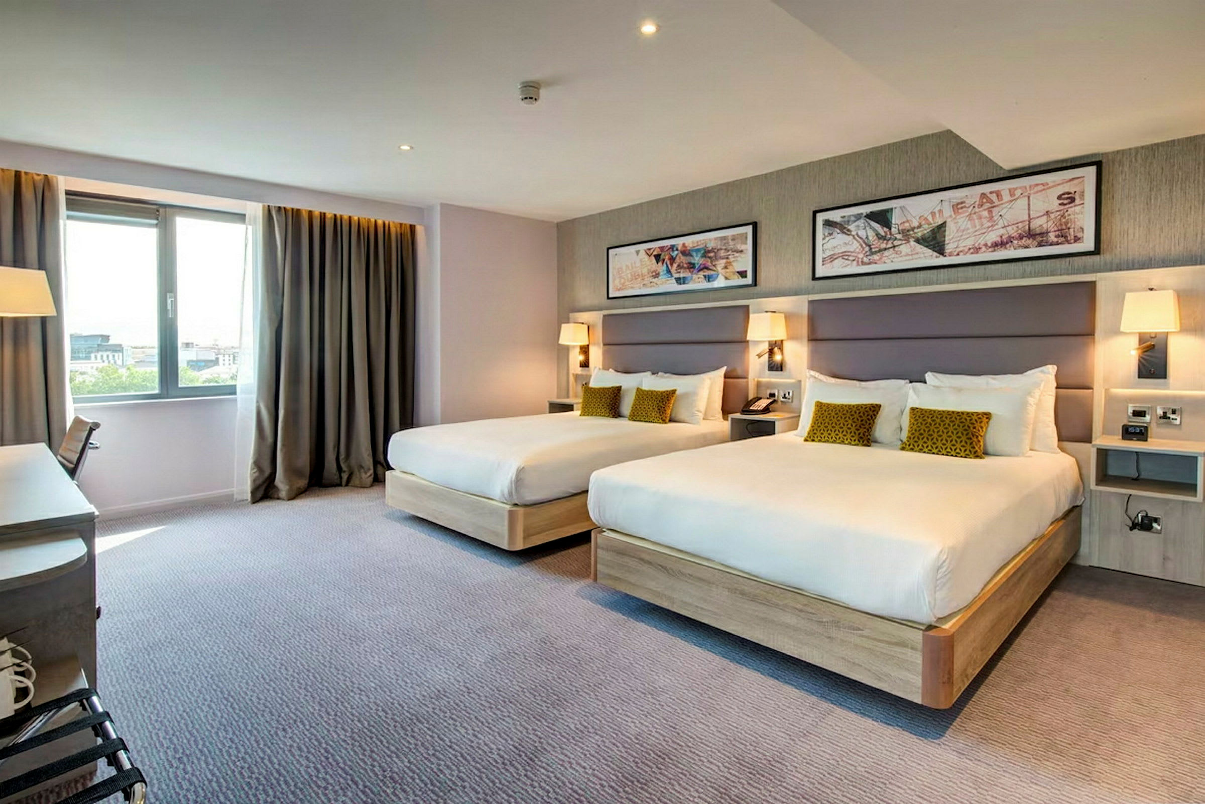 Två sängar med vita sängkläder och prydnadskuddar i ett hotellrum, med väggkonst ovanför och fönster vid sidan. (AI-genererad text)