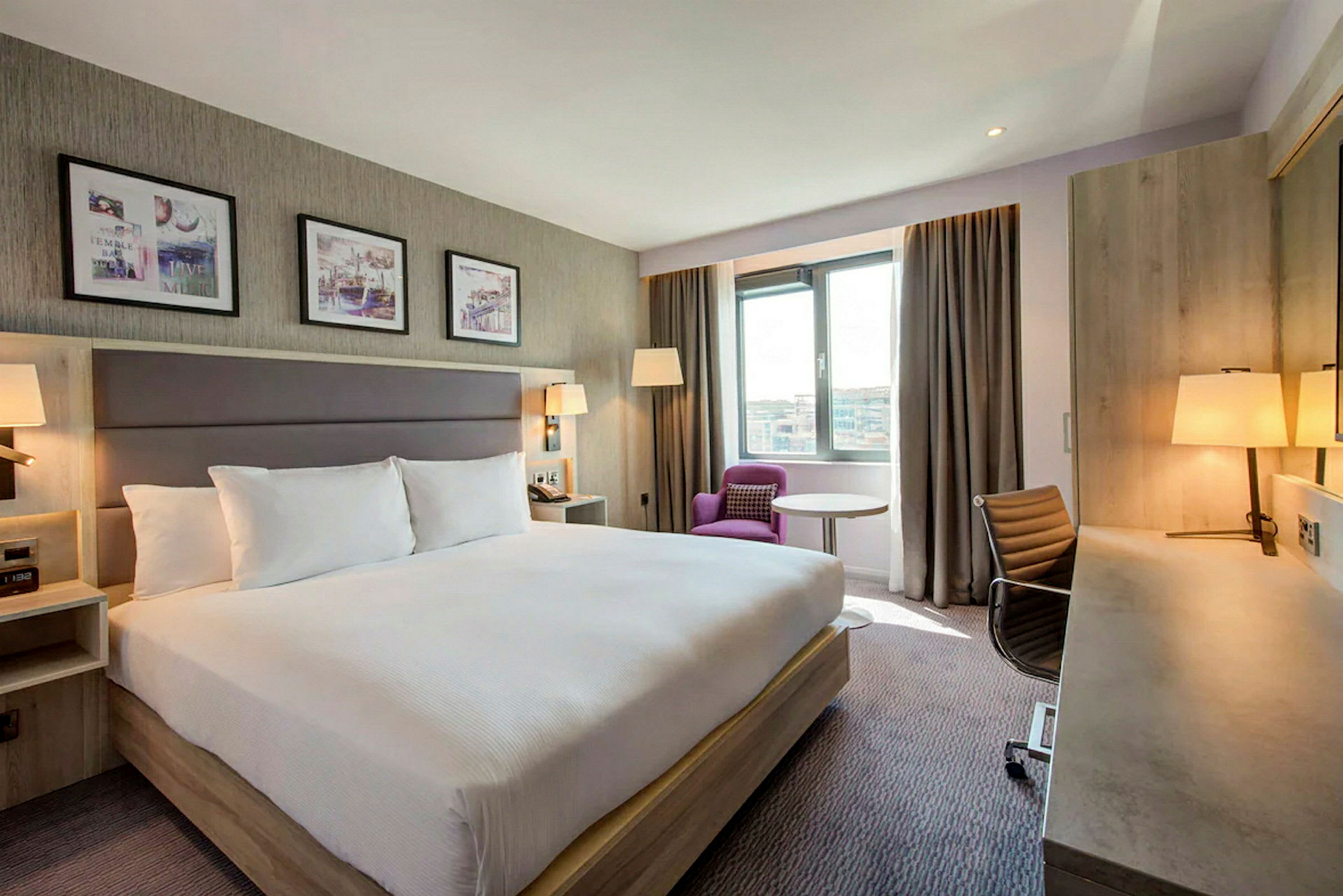 Ett hotellrum med en stor dubbelsäng, skrivbord, stol och en fåtölj vid fönstret. (AI-genererad text)