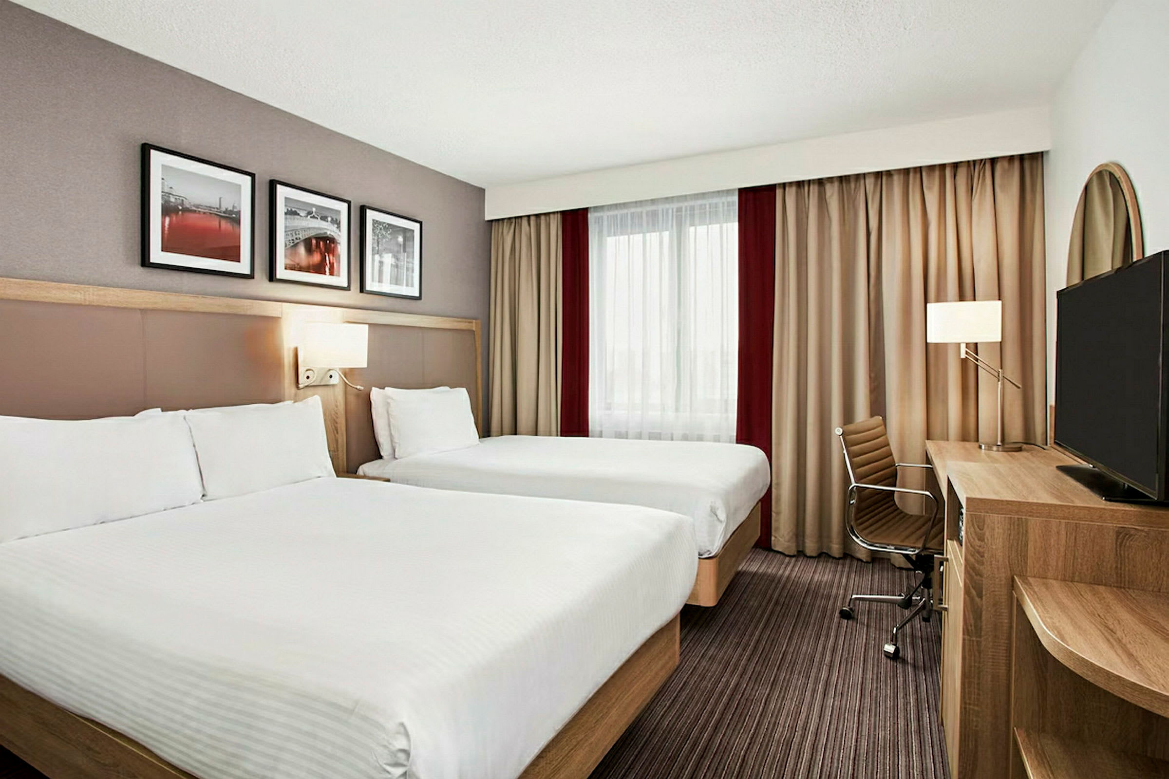 Ett hotellrum med två separata sängar, väggdekorationer, ett skrivbord, en stol och en TV. (AI-genererad text)