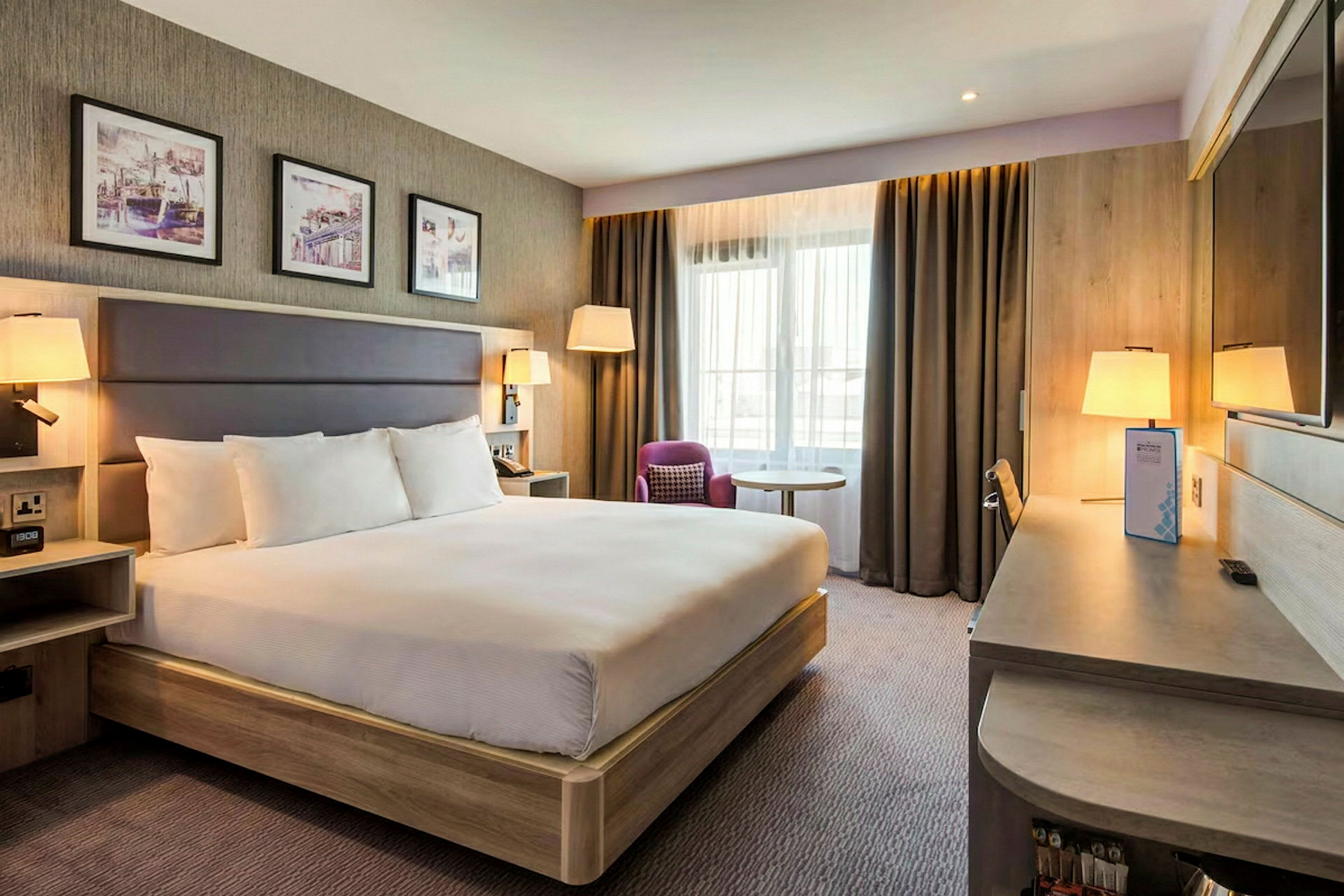 Hotellrum med en stor säng, skrivbord, stol och fönster med gardiner. (AI-genererad text)