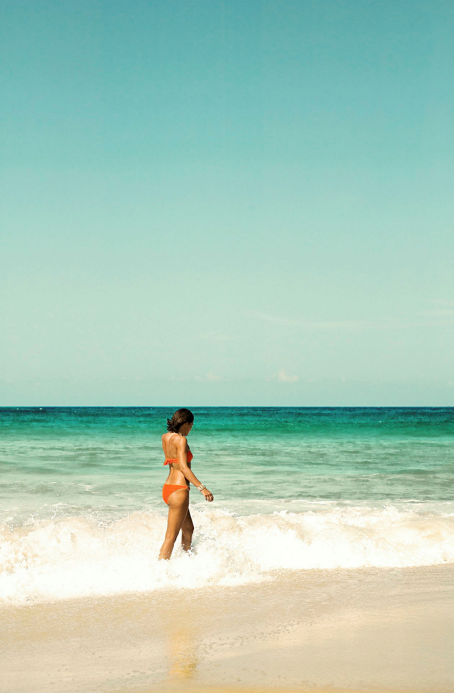 En kvinna i orange bikini går vid stranden nära vågorna. (AI-genererad text)