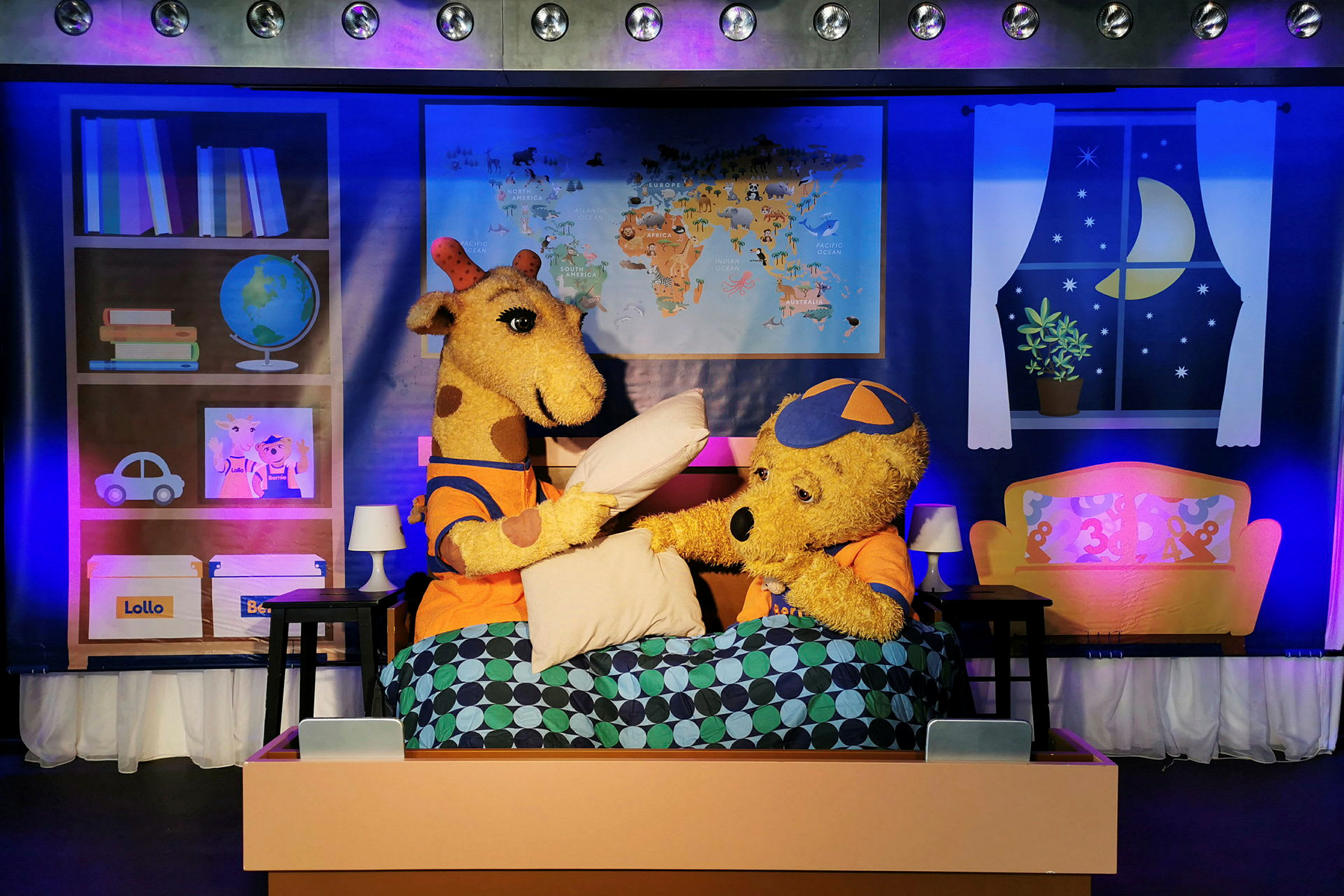 Två tecknade figurerdjur, en giraff och en björn, sitter i en säng med täcke och scenrekvisita synliga i bakgrunden. (AI-genererad text)