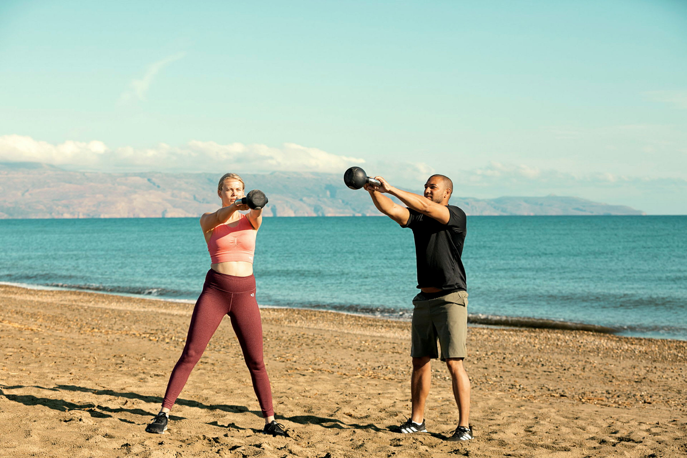 Två personer tränar med kettlebells på en sandstrand med havet i bakgrunden. (AI-genererad text)