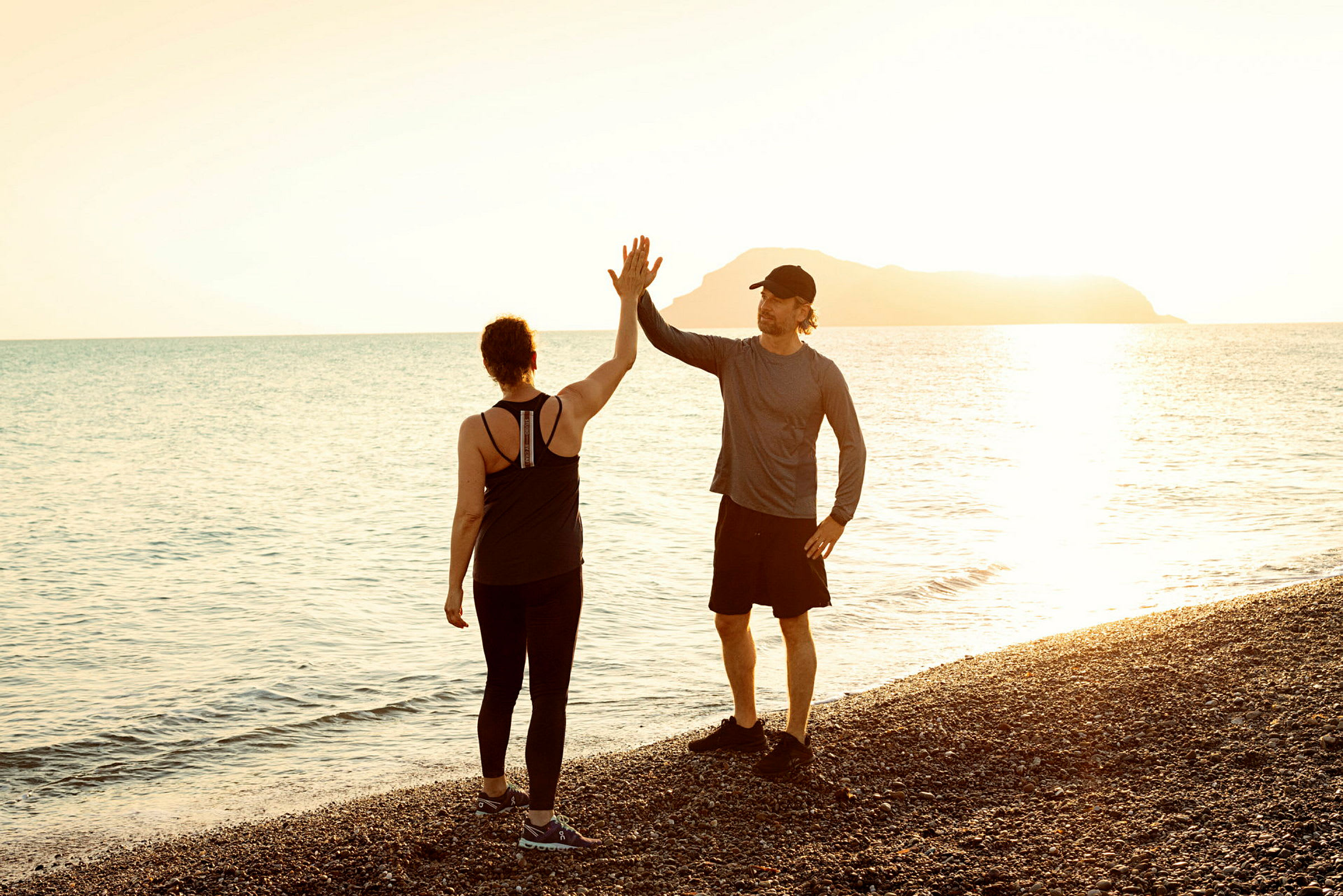 Två personer står på en stenstrand, ger varandra en high five under solnedgången över havet. (AI-genererad text)
