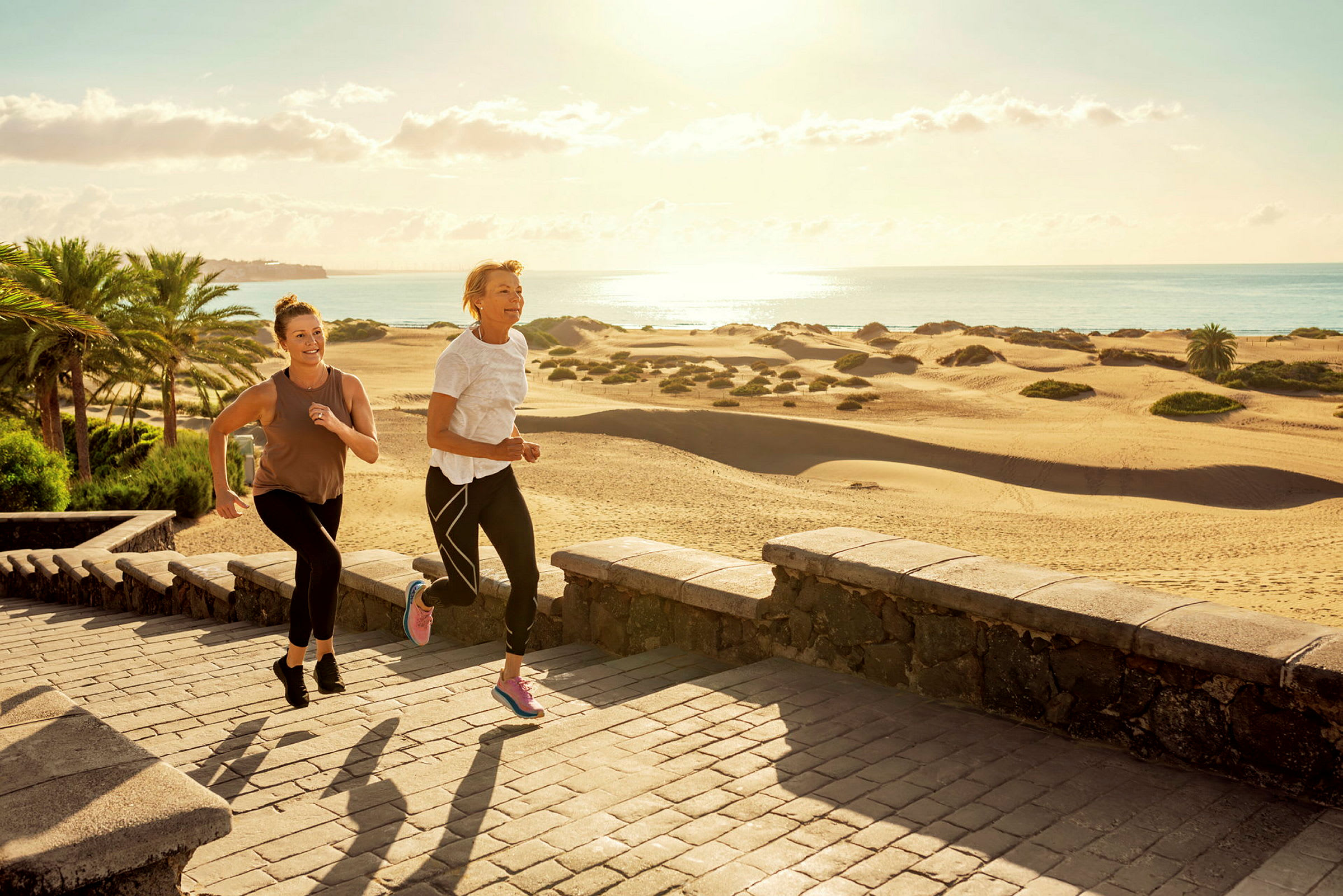 Två personer joggar på en stenlagd gångväg nära sanddynor och palmer vid kusten. (AI-genererad text)