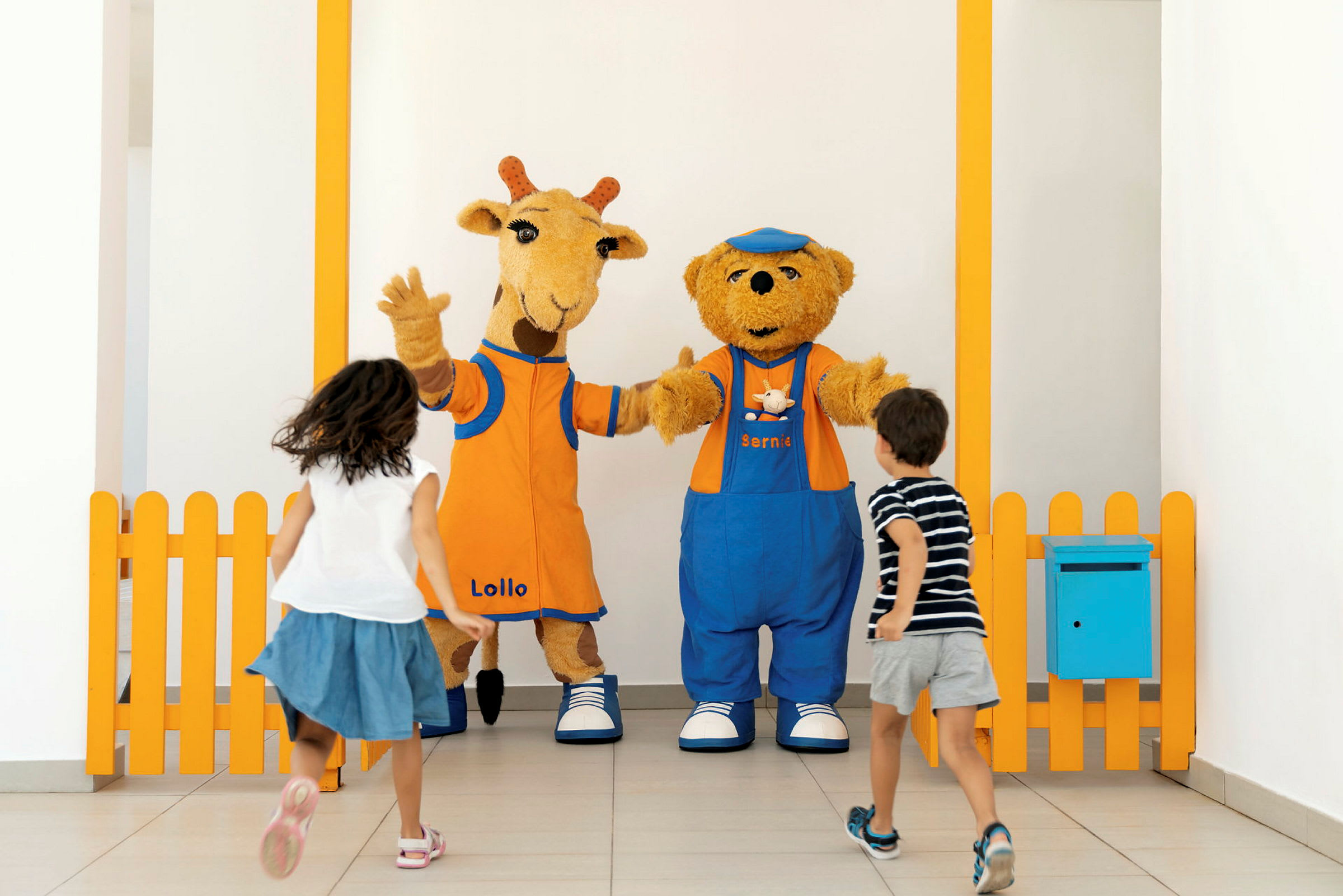 Två barn springer mot två maskotfigurer, en giraff och en björn, som står bredvid ett orange staket. (AI-genererad text)