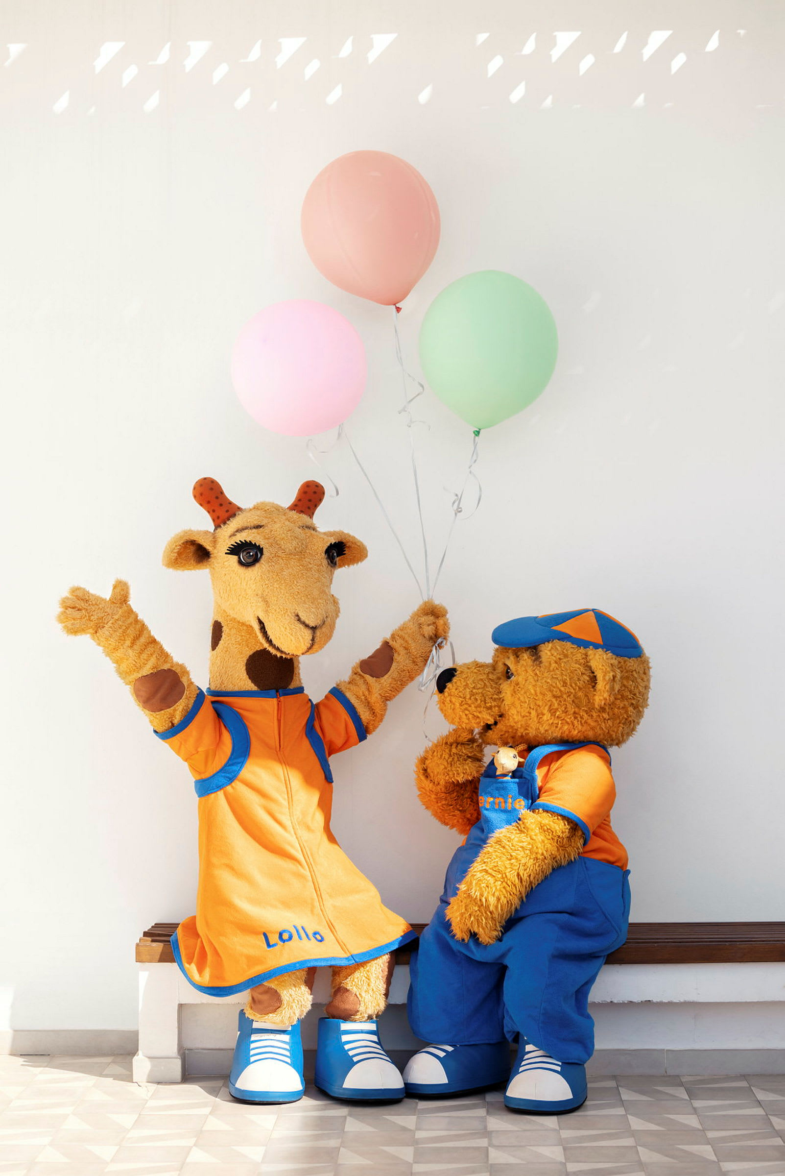 Två maskotar, en giraff och en björn, sitter på en bänk med ballonger. (AI-genererad text)
