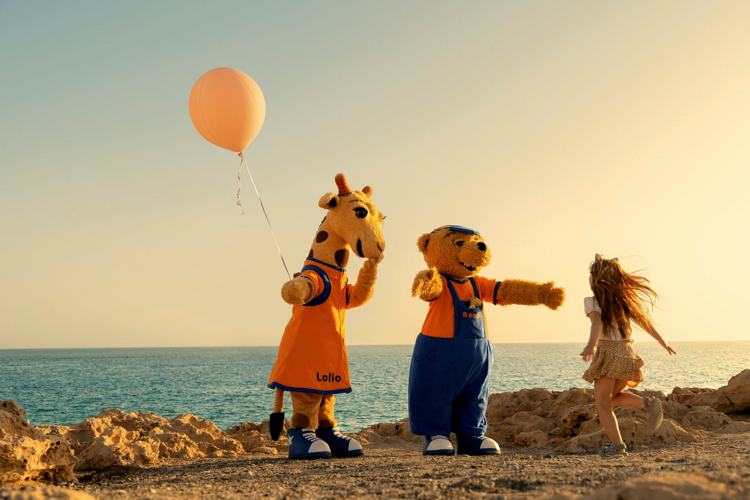 Två karaktärer klädda som en giraff och en björn står bredvid ett barn på en strand med havet i bakgrunden. Giraffen håller en orange ballong. (AI-genererad text)