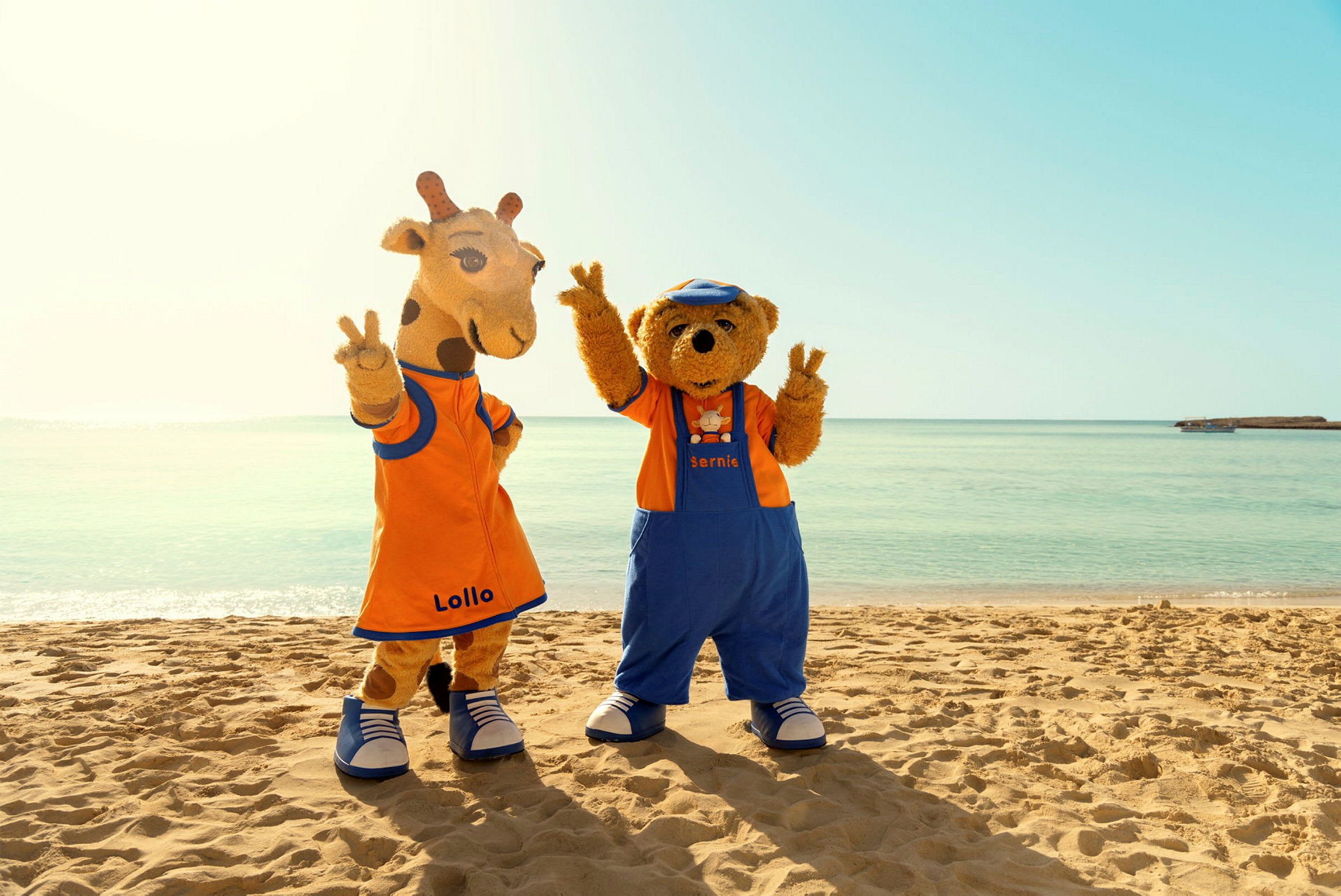 Två maskotkaraktärer med namnen Lollo och Bernie står på en strand framför klara havet. (AI-genererad text)