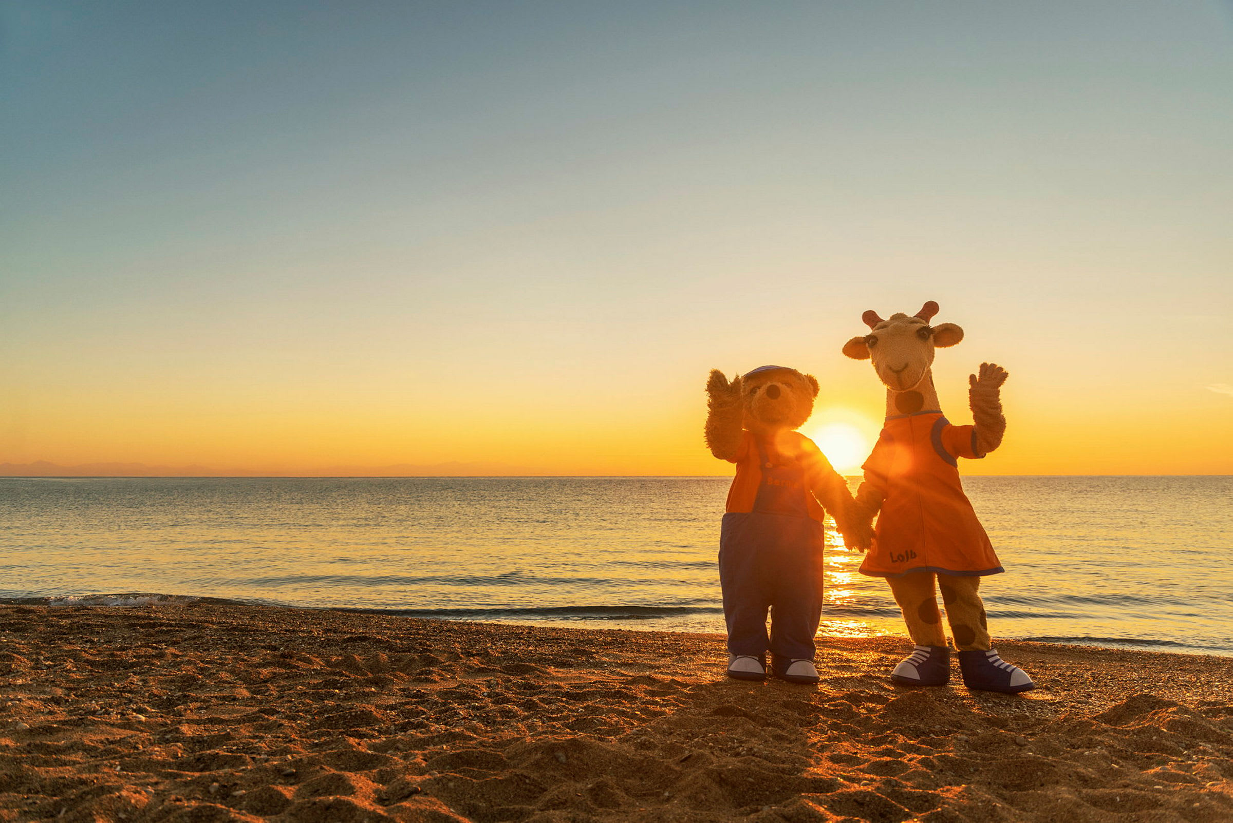 Två maskotar står på en strand framför en solnedgång, vänd mot kameran. (AI-genererad text)