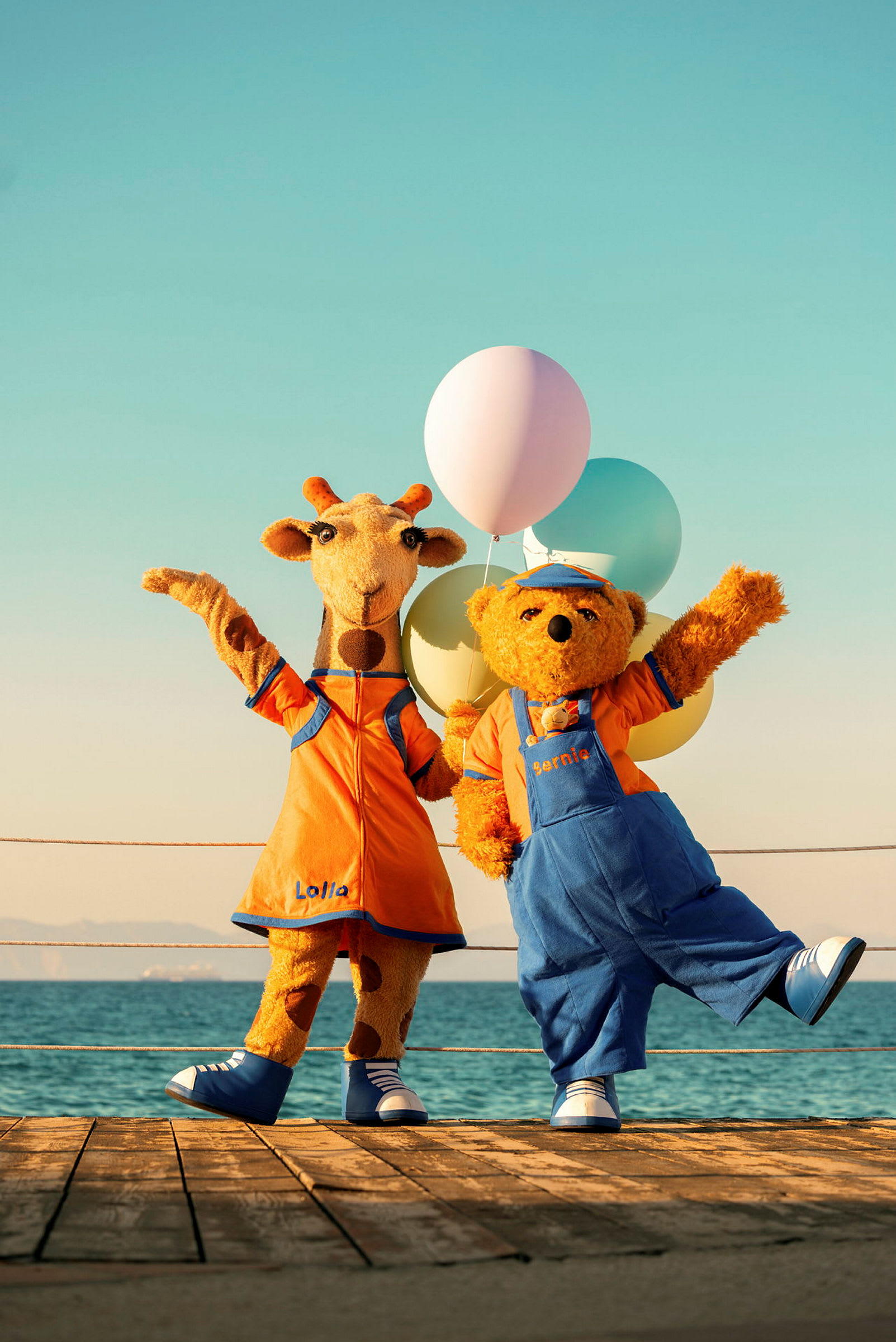 Två figurer, en giraff och en björn, klädda i färgglada dräkter står på en brygga med ballonger. (AI-genererad text)