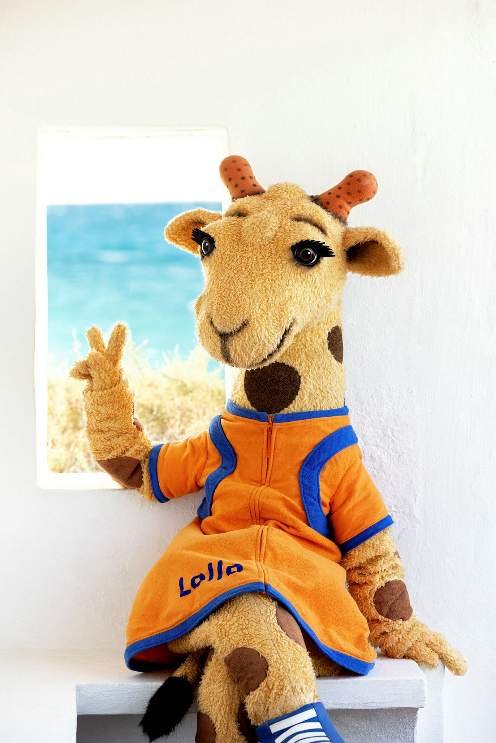 En mjukisdocka föreställande en giraff sitter och håller upp två fingrar. I bakgrunden syns havet. (AI-genererad text)