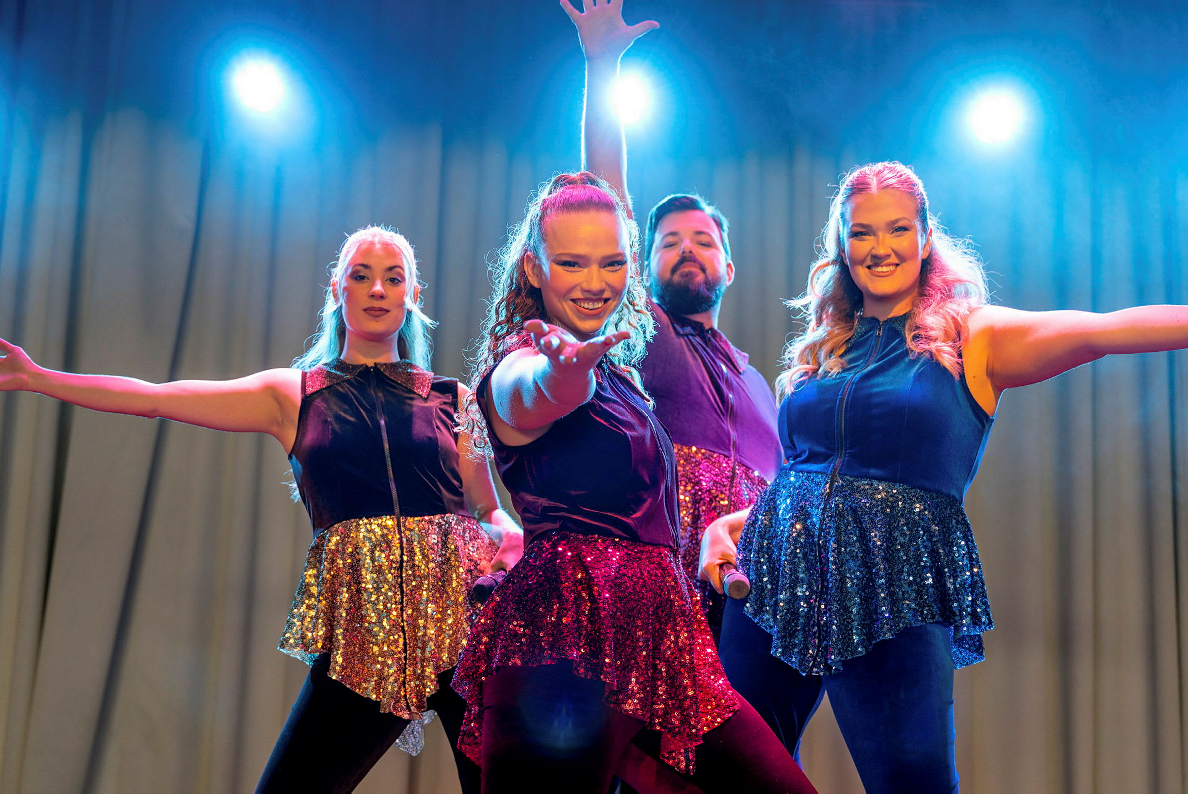 Fyra personer i glittrande scenkläder poserar och uppträder på en upplyst scen. (AI-genererad text)
