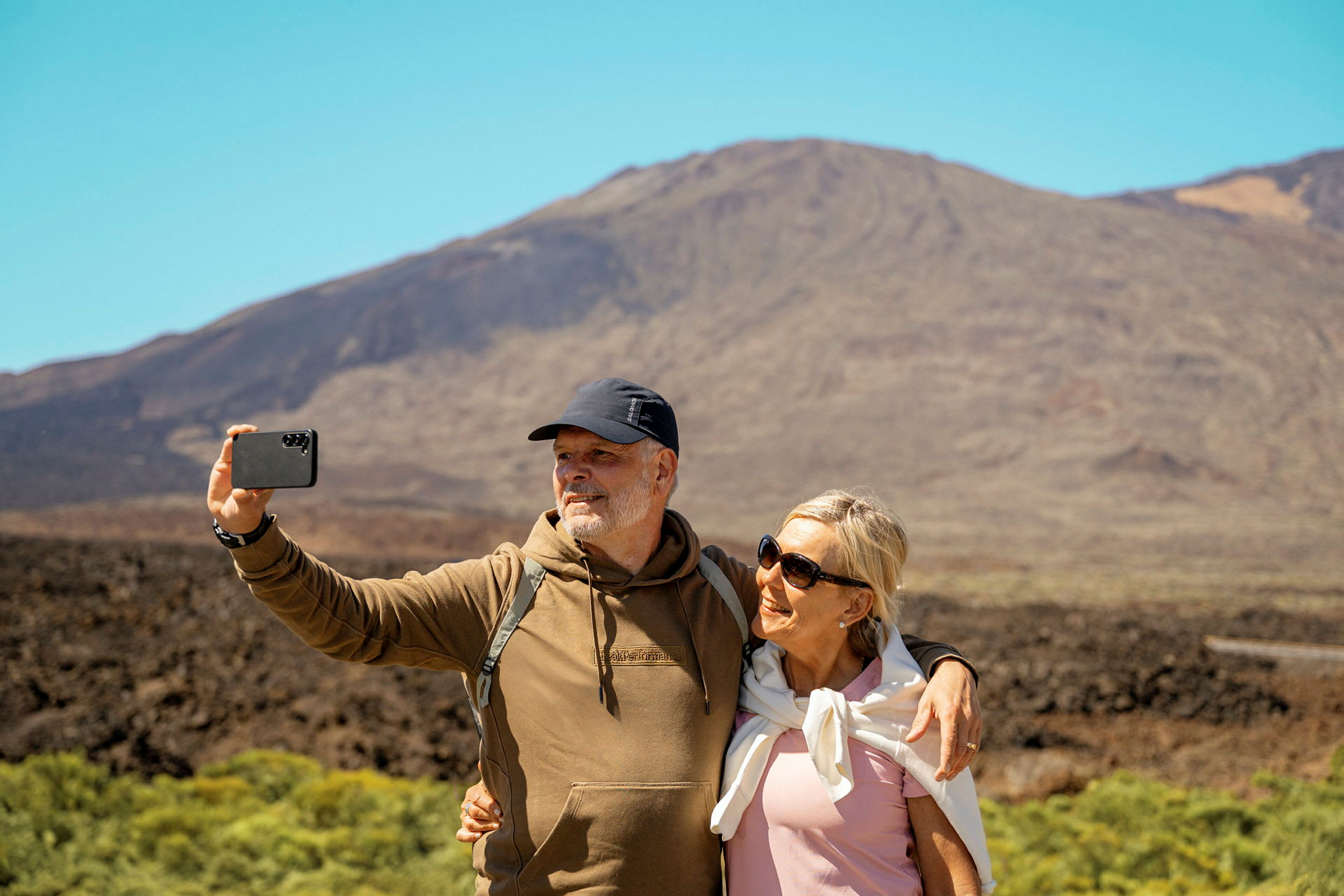 Två personer tar en selfie framför ett berg i soligt väder. (AI-genererad text)