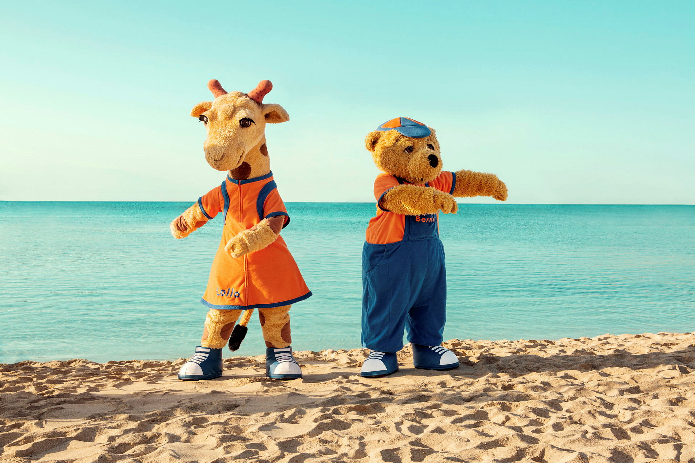 Två maskotar, en giraff och en björn, står på en strand med havet i bakgrunden. (AI-genererad text)
