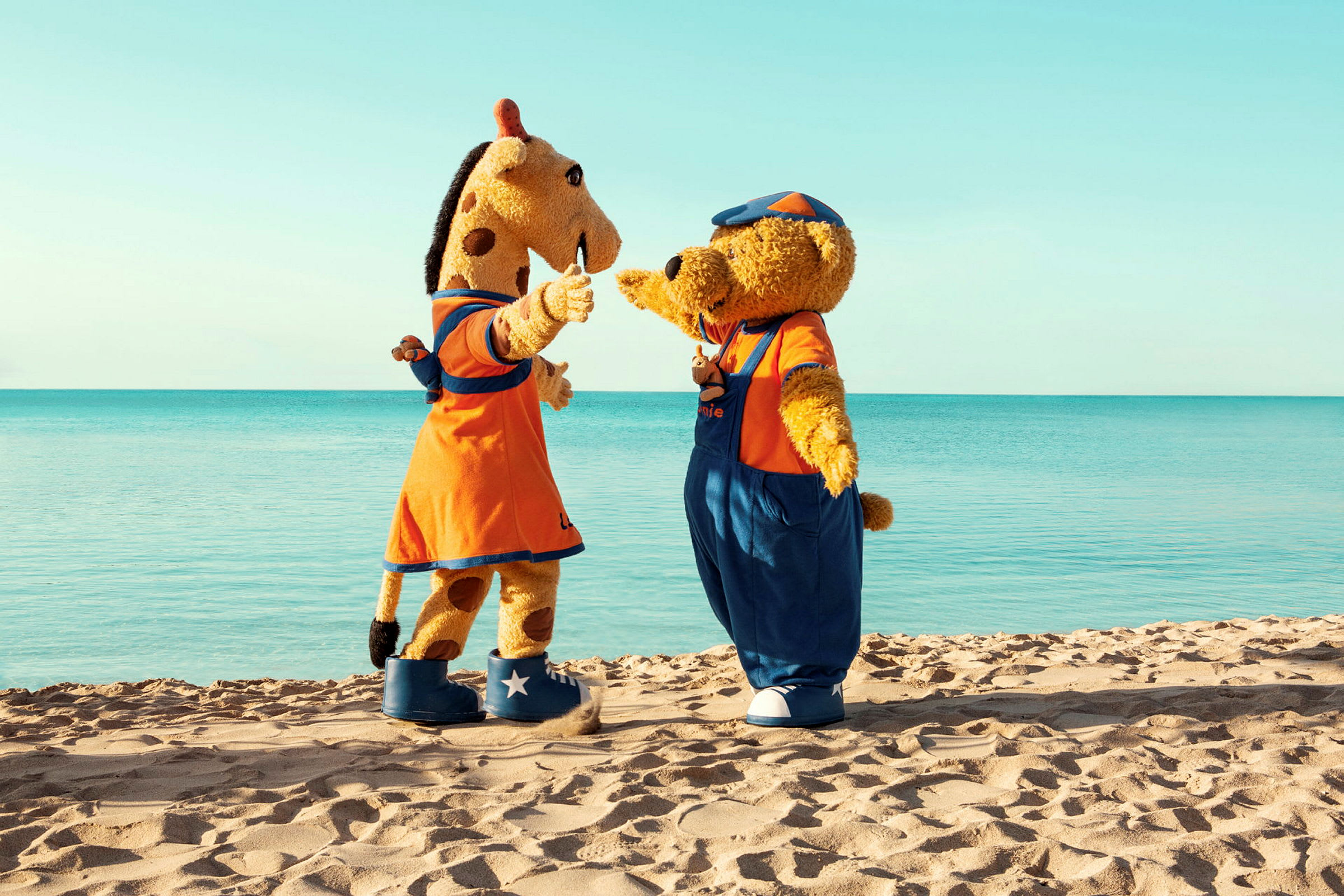 Två maskotar, en giraff och en björn, står på en sandstrand med havet i bakgrunden. (AI-genererad text)