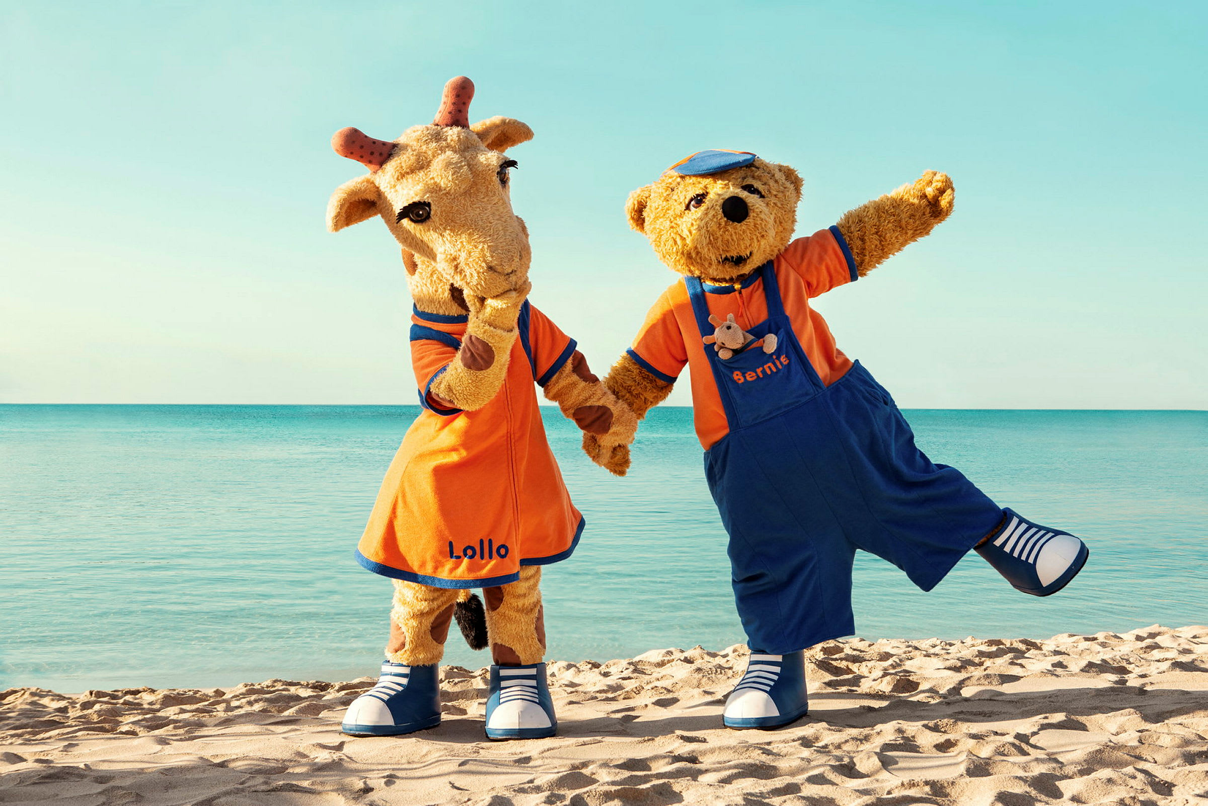 Maskotarna Lollo och Bernie står på en sandstrand framför havet. (AI-genererad text)