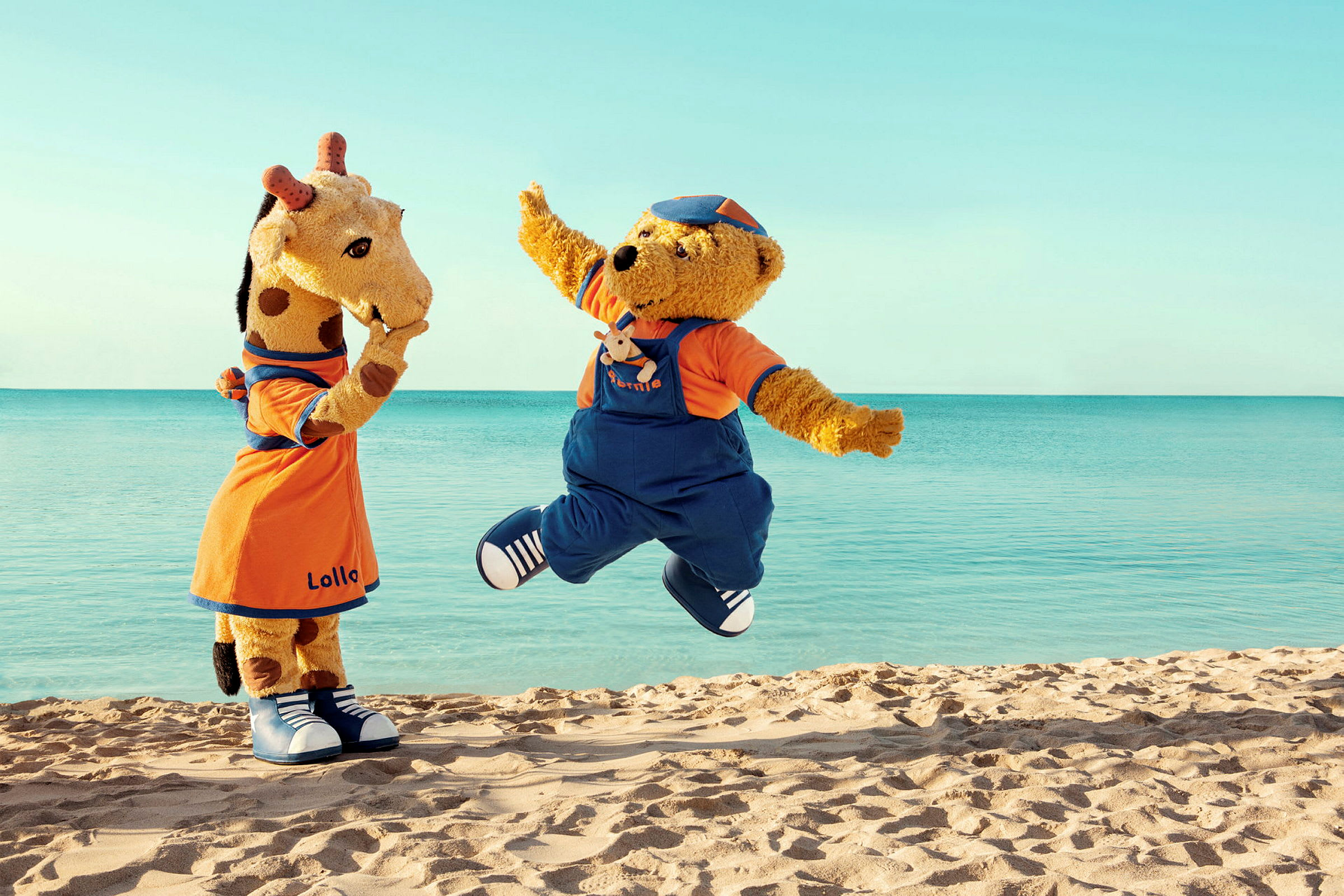 Två maskotfigurer, en giraff och en björn, står på en strand framför havet. (AI-genererad text)