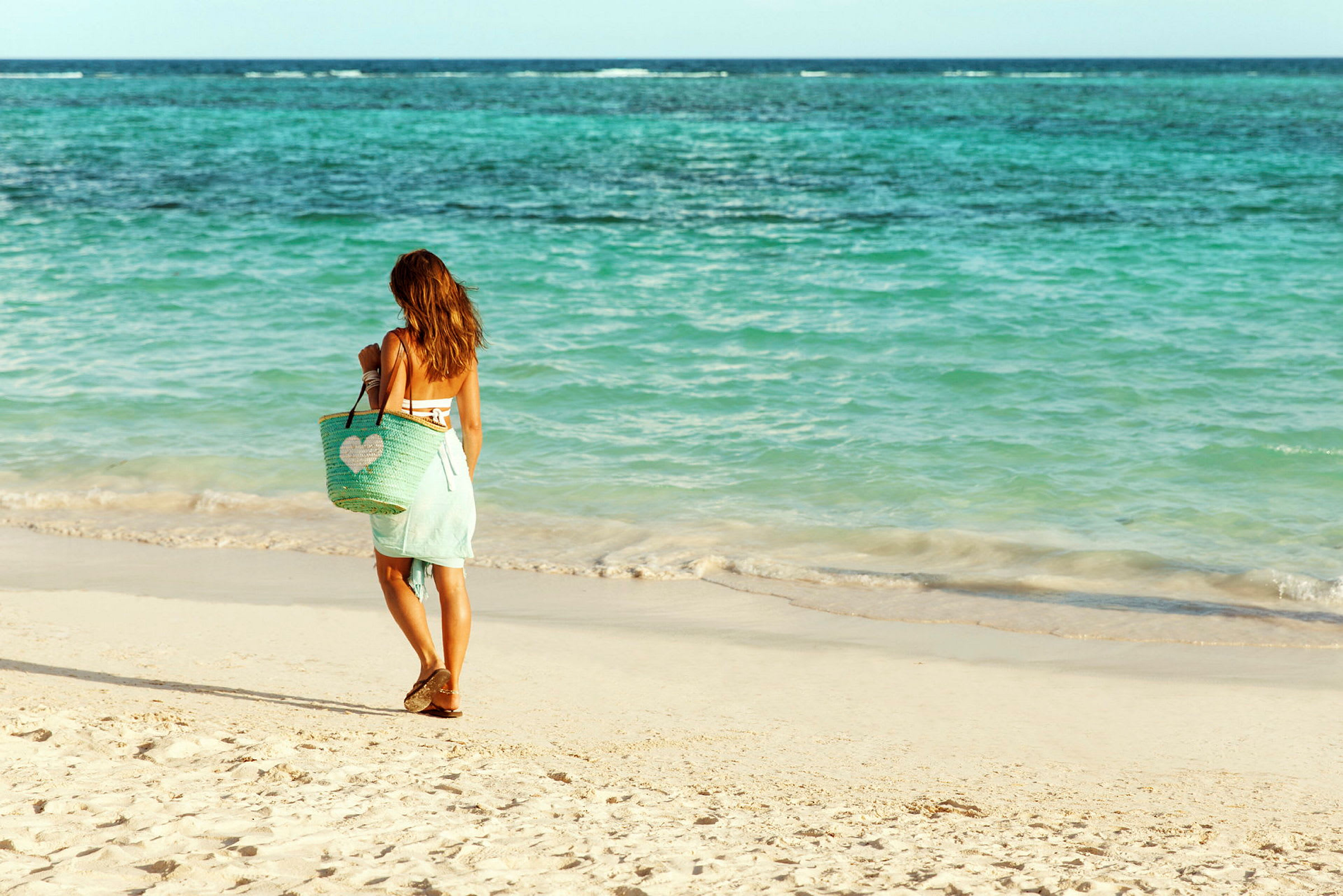 En person går på en sandstrand nära havet, bärande en strandväska. (AI-genererad text)
