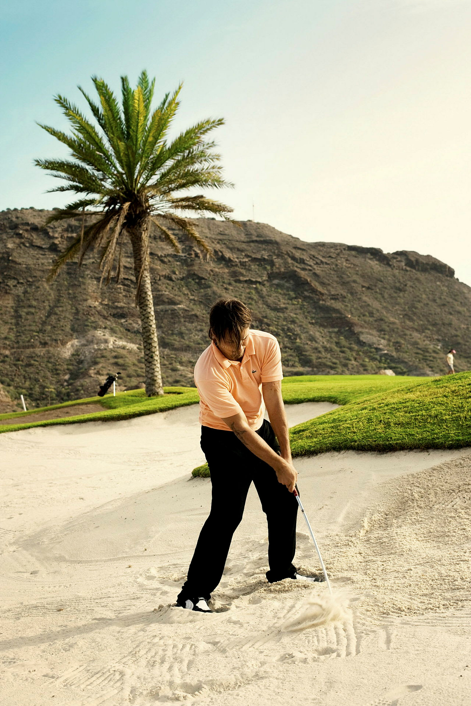 En person spelar golf i en sandbunker med en golfklubba. I bakgrunden syns en palm och berg. (AI-genererad text)