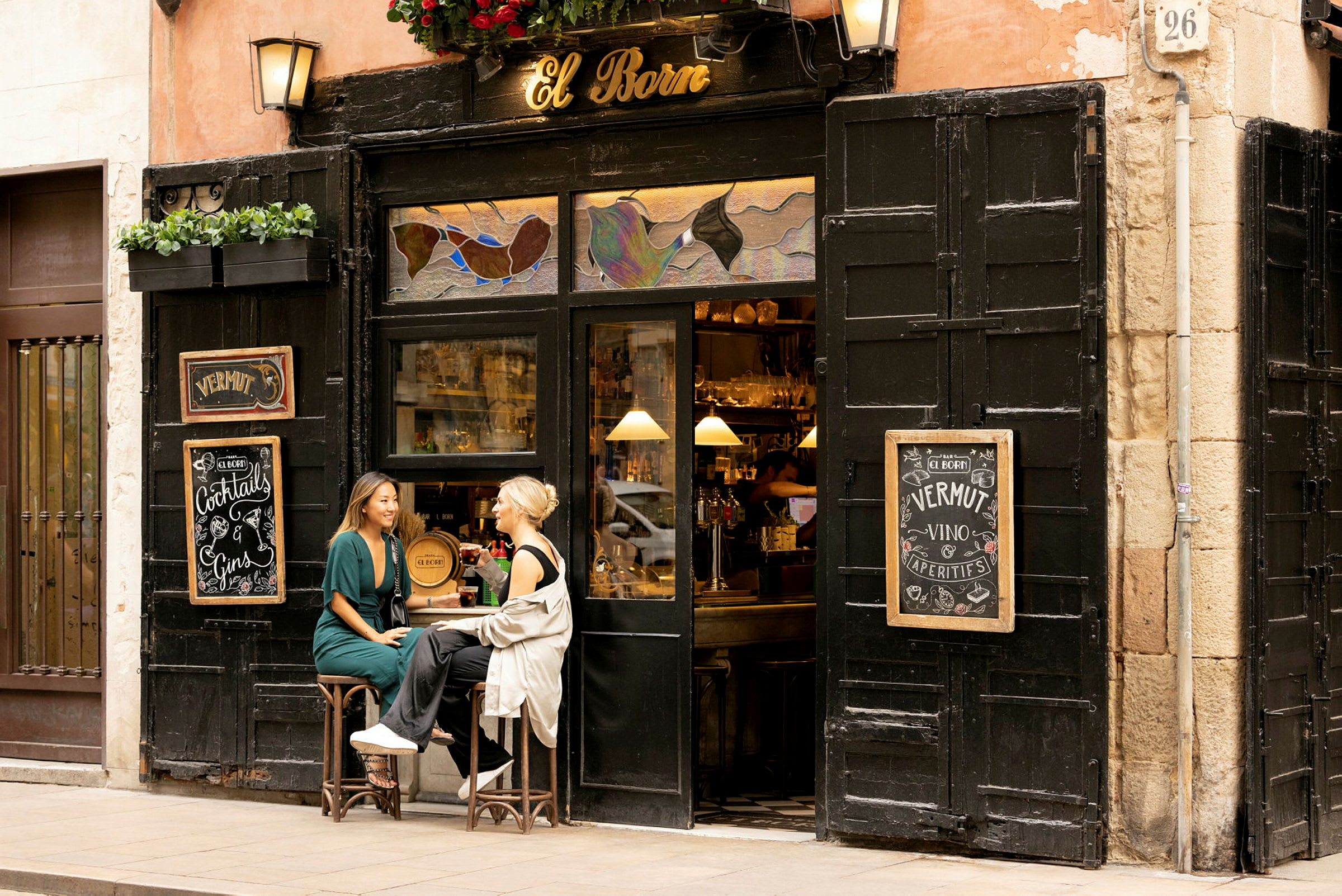 Två personer sitter på pallar utanför en liten bar med skylten 'El Born'. (AI-genererad text)
