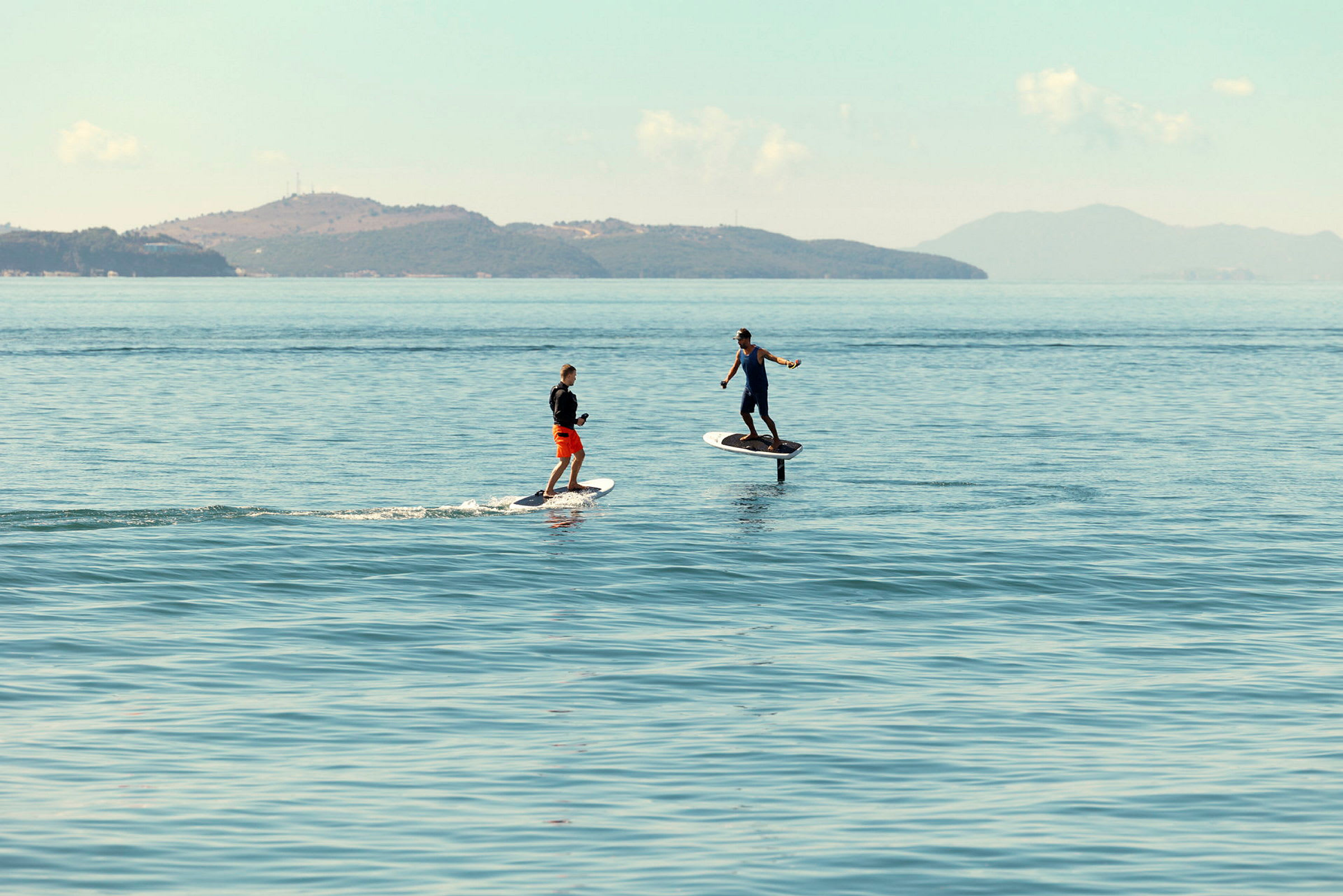 Två personer står på elektriska surfbrädor på en lugn havsytan med berg i bakgrunden. (AI-genererad text)