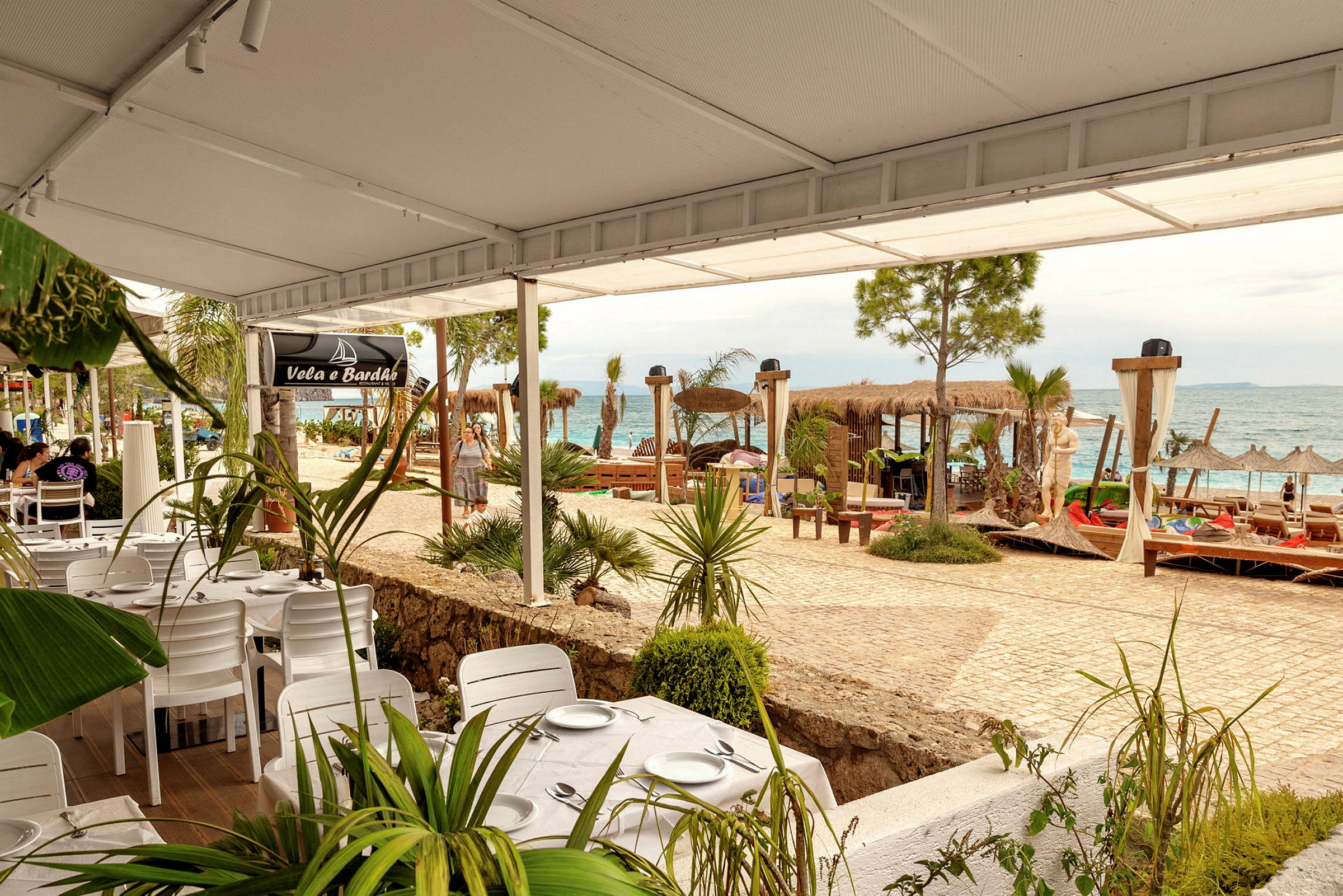 Strandnära restaurang med vita bord och stolar, havsutsikt och solstolar. (AI-genererad text)