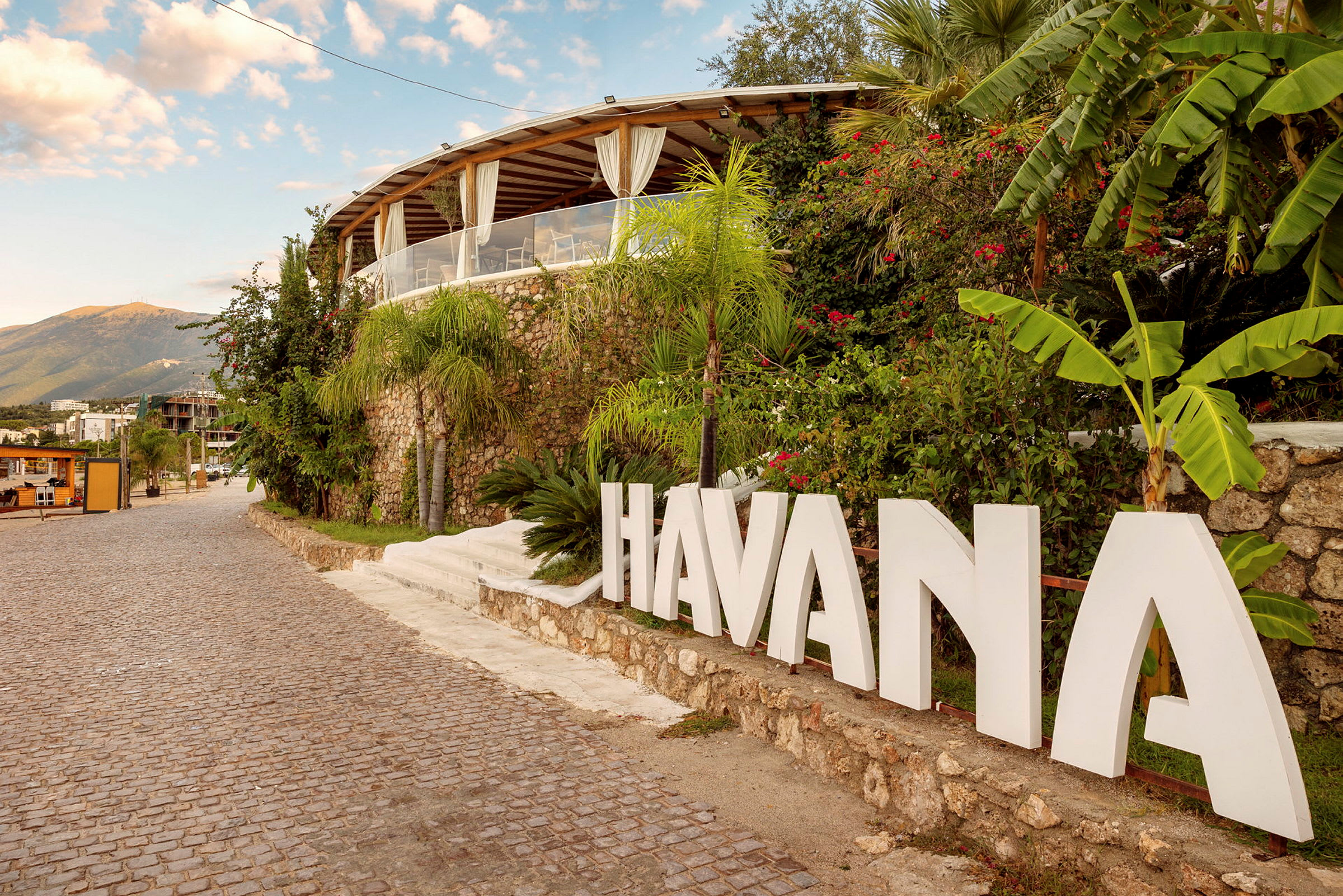Skriven text med 'HAVANA' vid en tropisk byggnad bredvid kullerstensväg. (AI-genererad text)