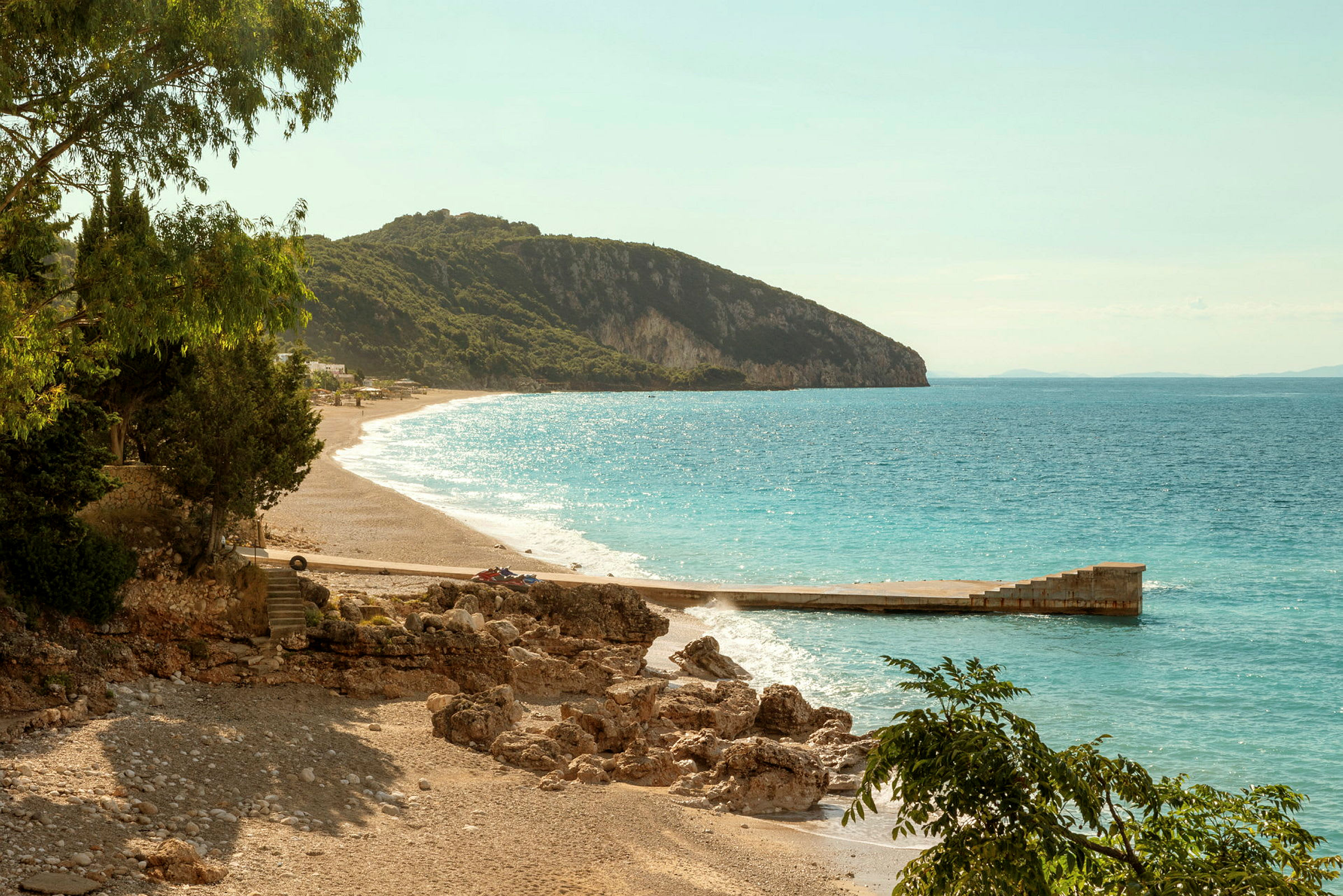 Strand med sand och klippor bredvid havet med en träbrygga och skogsklädda berg i bakgrunden. (AI-genererad text)