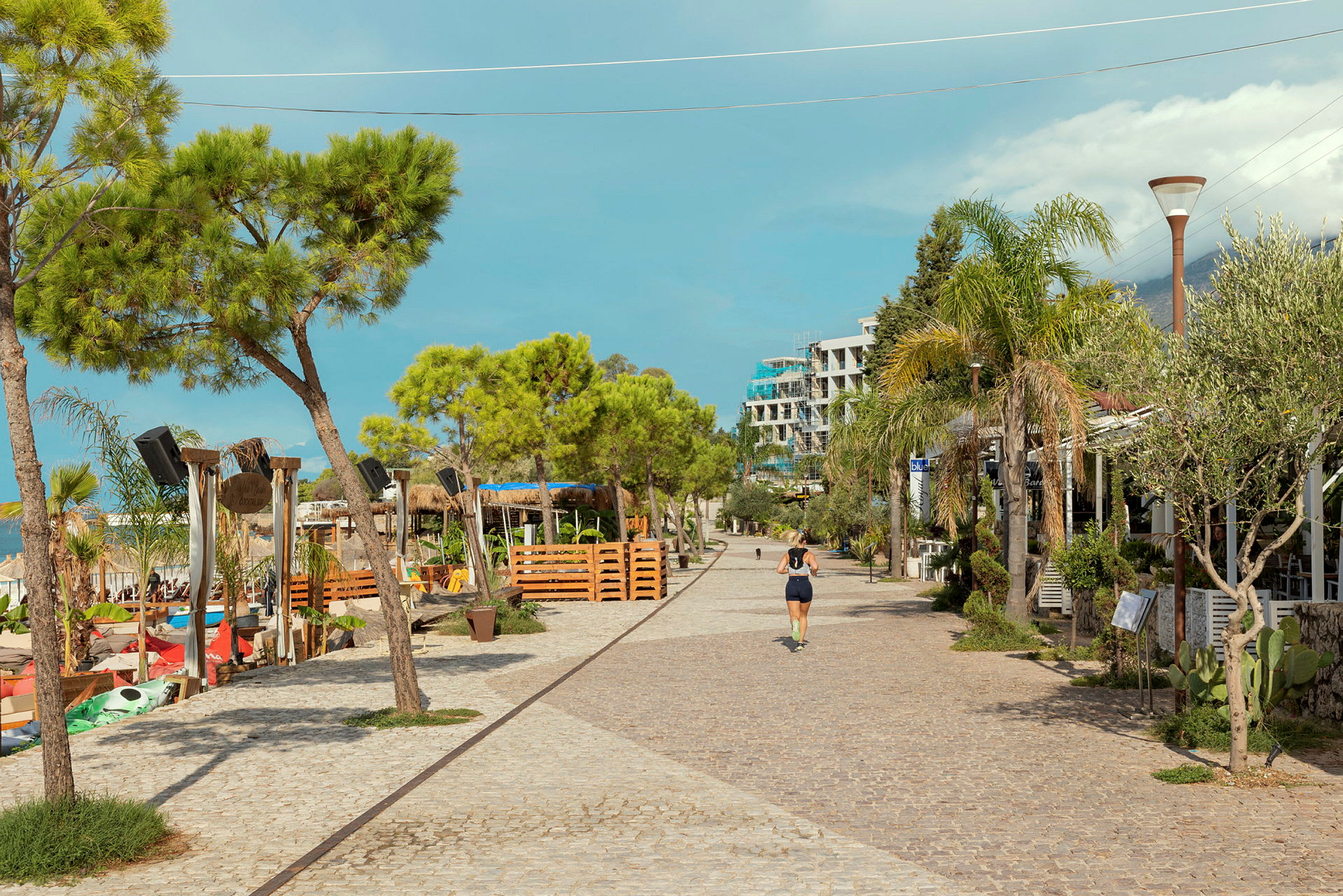 En stenlagd promenadväg kantad av träd, restauranger och ett strandområde. (AI-genererad text)