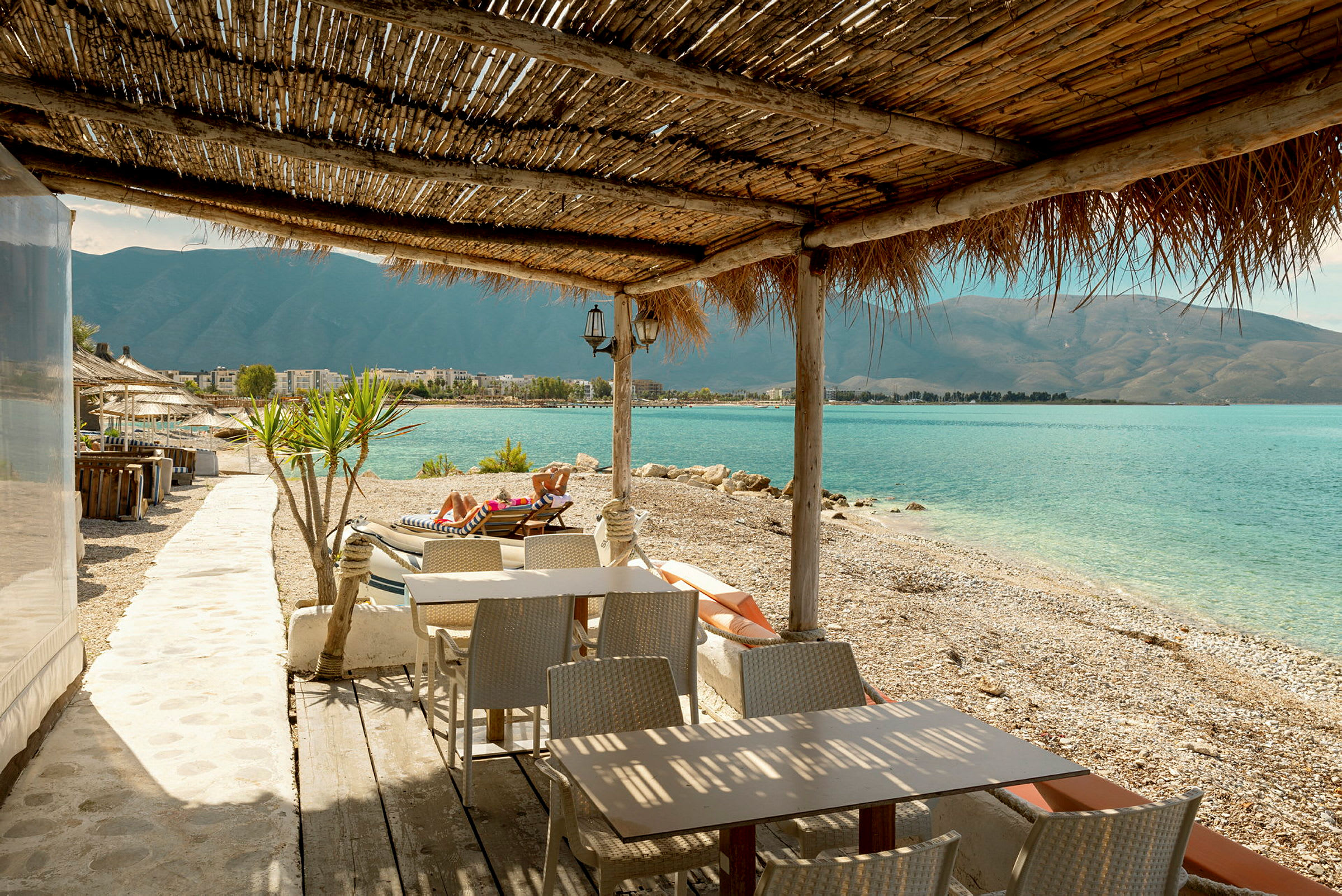 En restaurang vid en stenstrand med utsikt över havet och berg. (AI-genererad text)