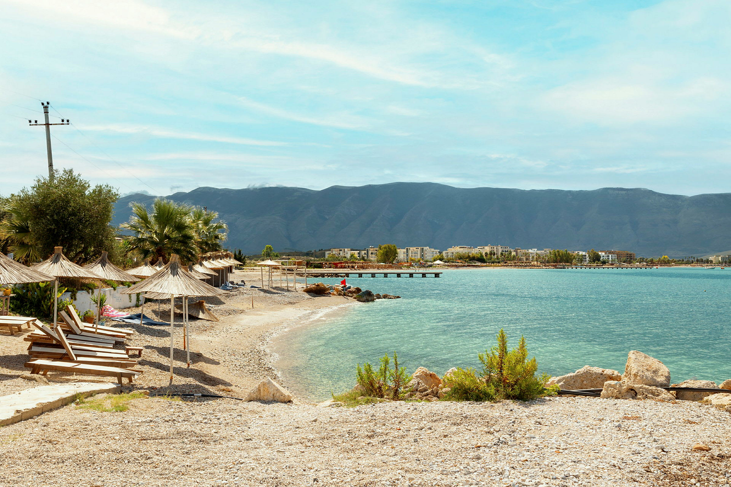 Strand med solstolar och parasoller vid klart vatten och fjärran berg. (AI-genererad text)