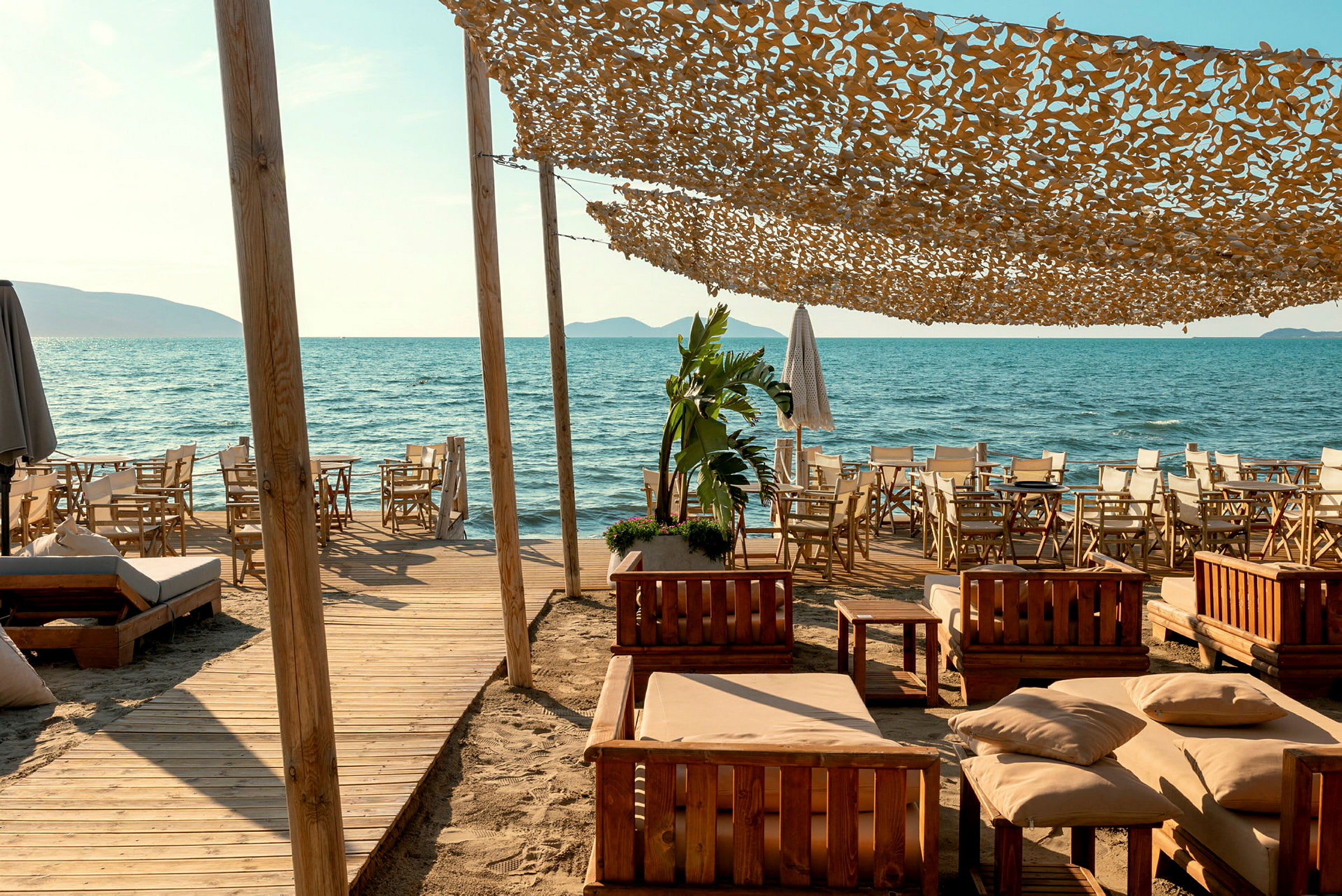 En strand med en uteservering, möbler i trä och bekväma sittplatser, samt ett tygsegel som skyddar mot solen. (AI-genererad text)