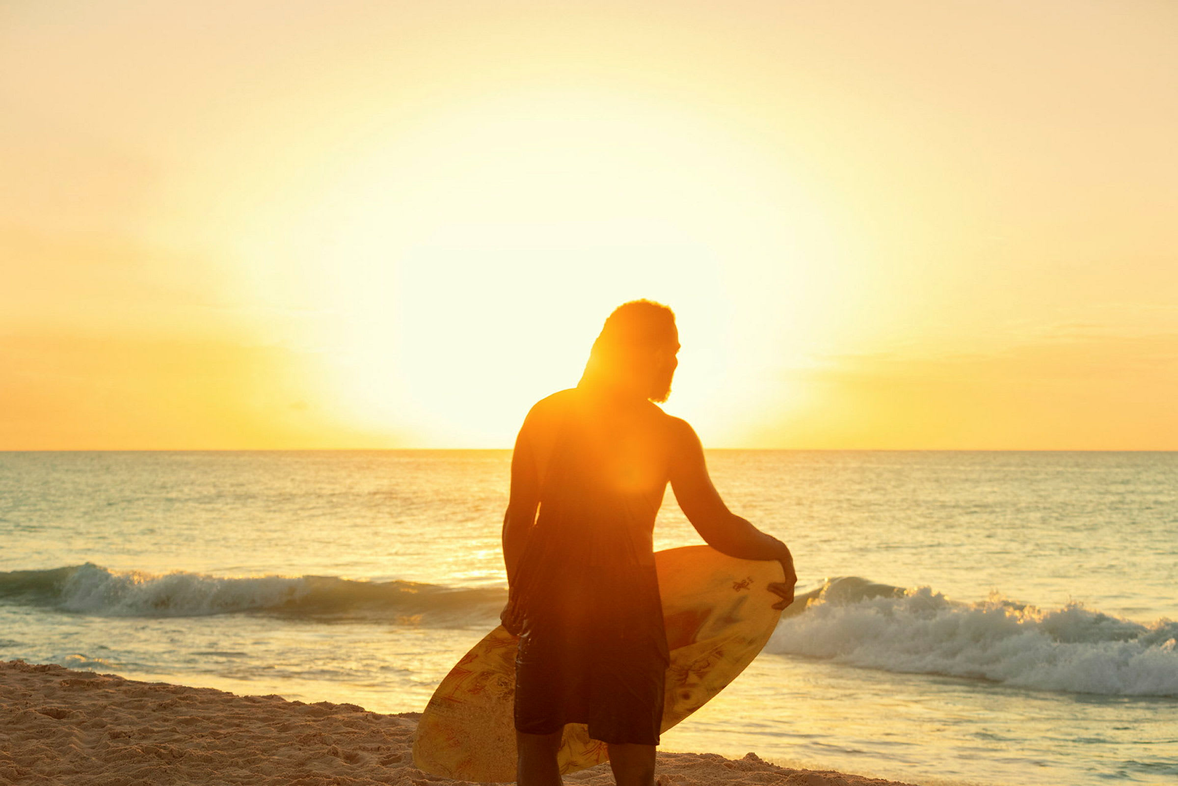 En person står på stranden vid havet, håller en surfbräda och solen lyser starkt i bakgrunden. (AI-genererad text)
