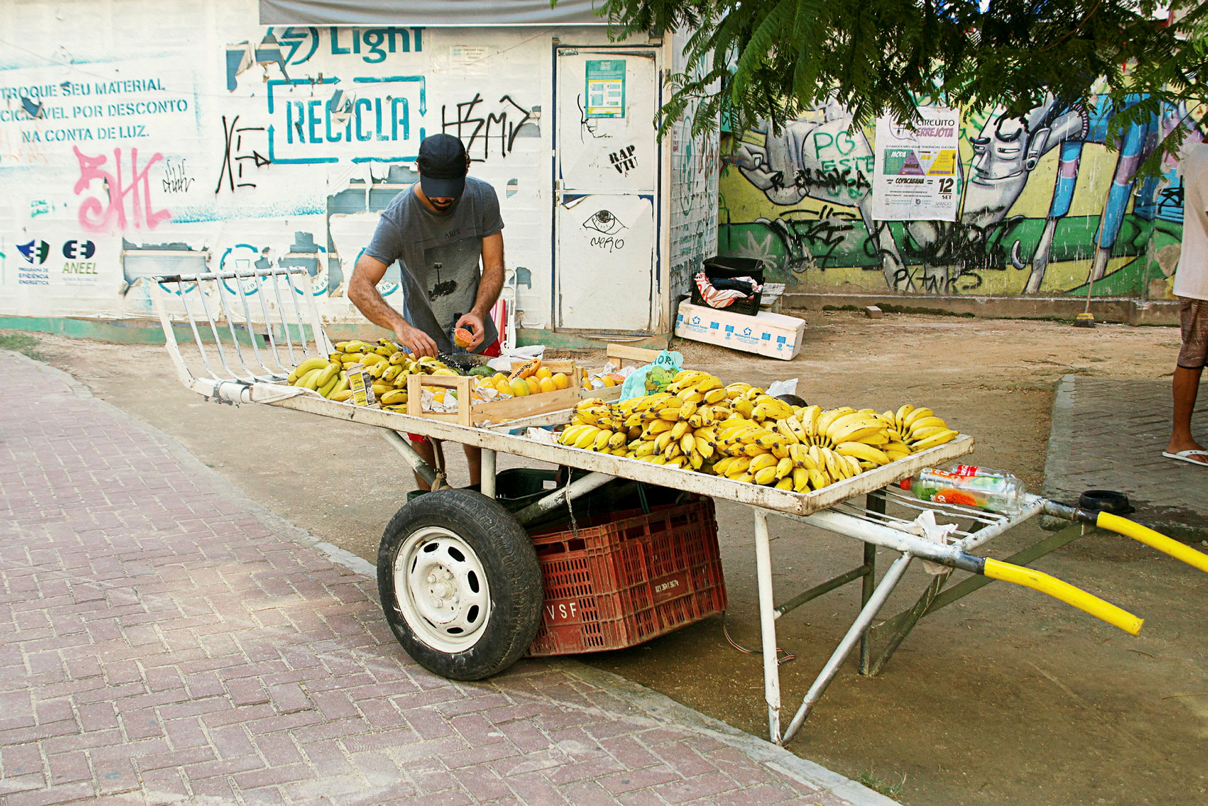 En man ordnar en fruktvagn fylld med bananer och andra frukter på en trottoar. (AI-genererad text)