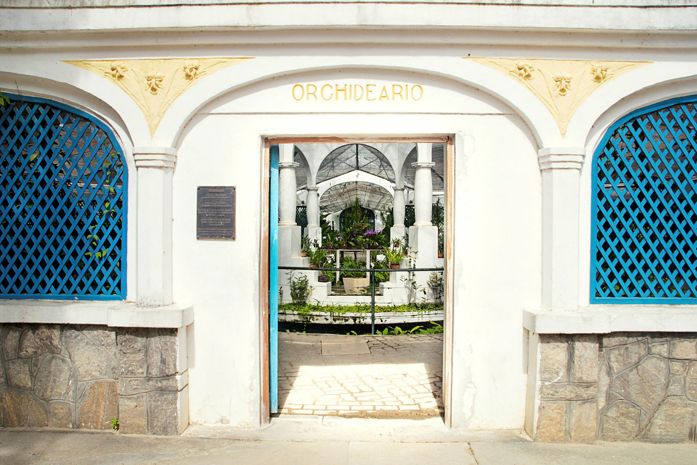 En vit byggnad med en veranda med bågar och texten "Orchideario" ovanför entrén som leder till en trädgård. (AI-genererad text)