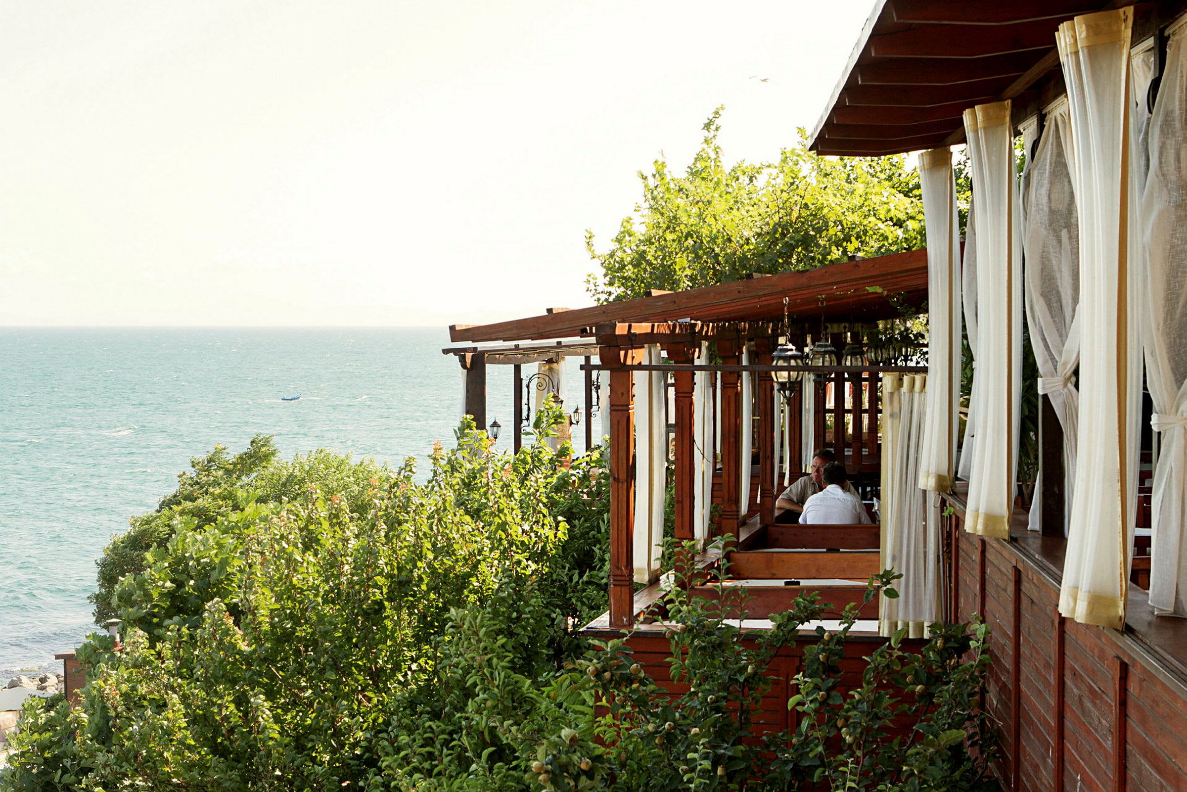 En restaurang med träpergola och vita draperier, omgiven av grönska, nära havet. (AI-genererad text)