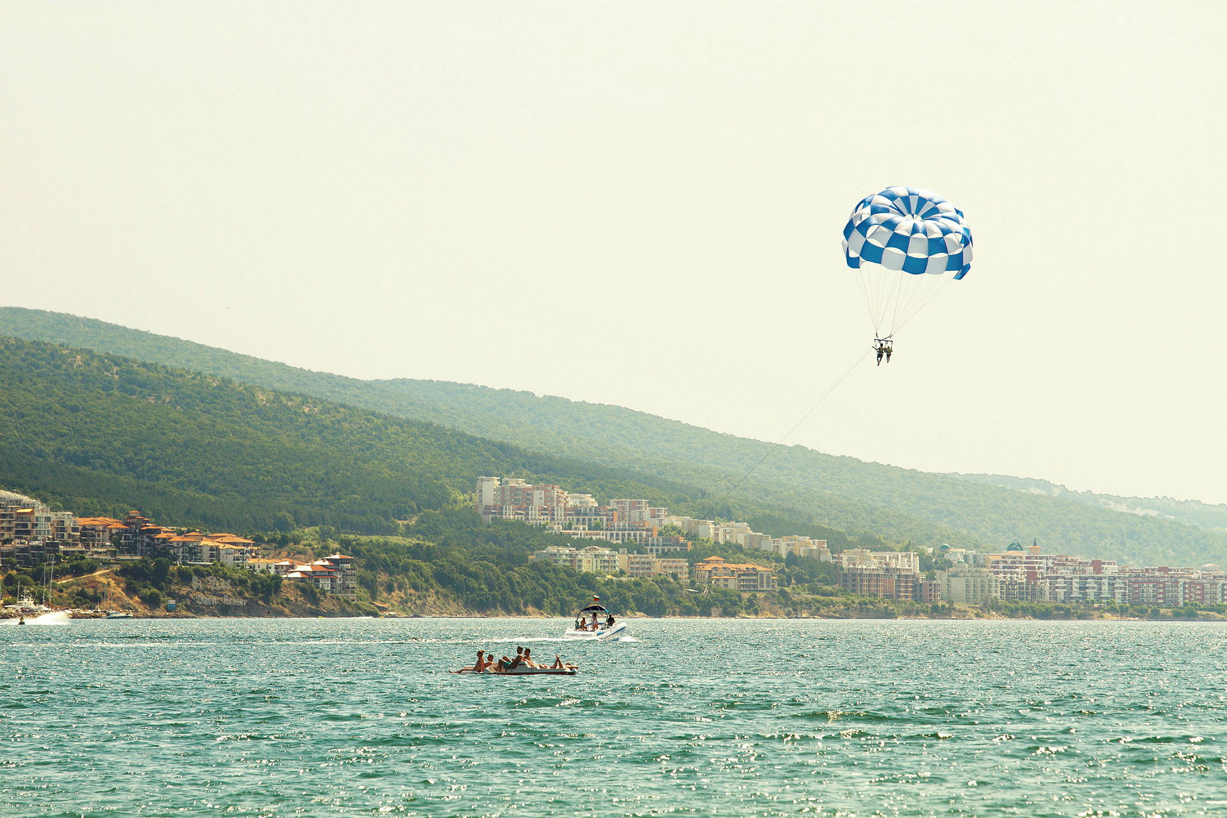Personer som parasailar med ett blåvitt fallskärm över vatten vid en kust. (AI-genererad text)