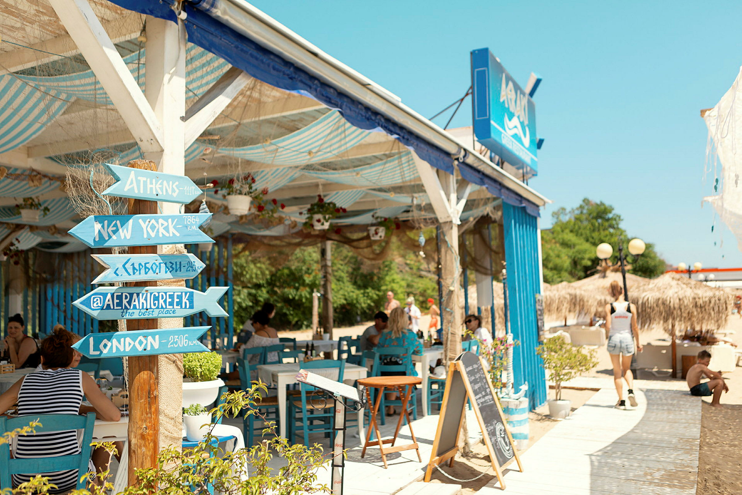En strandnära restaurang med blå möbler och en träskylt med destinationsnamn. (AI-genererad text)