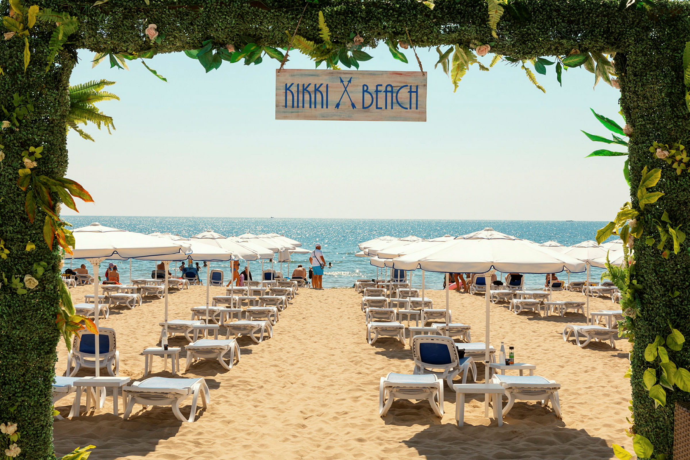 Solstolar och vita parasoller uppställda på en sandstrand med havet i bakgrunden, samt en skylt med texten "Kikki Beach" ovanför. (AI-genererad text)