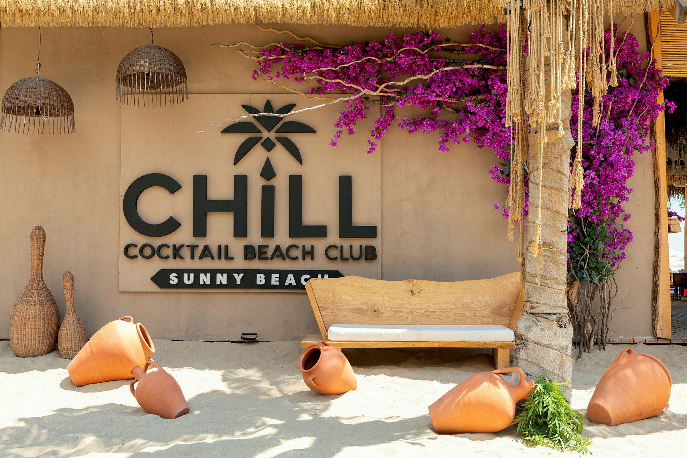 En strandbar med en skylt som säger 'Chill Cocktail Beach Club Sunny Beach', omgiven av terrakottakrukor och färgglada blommor. (AI-genererad text)