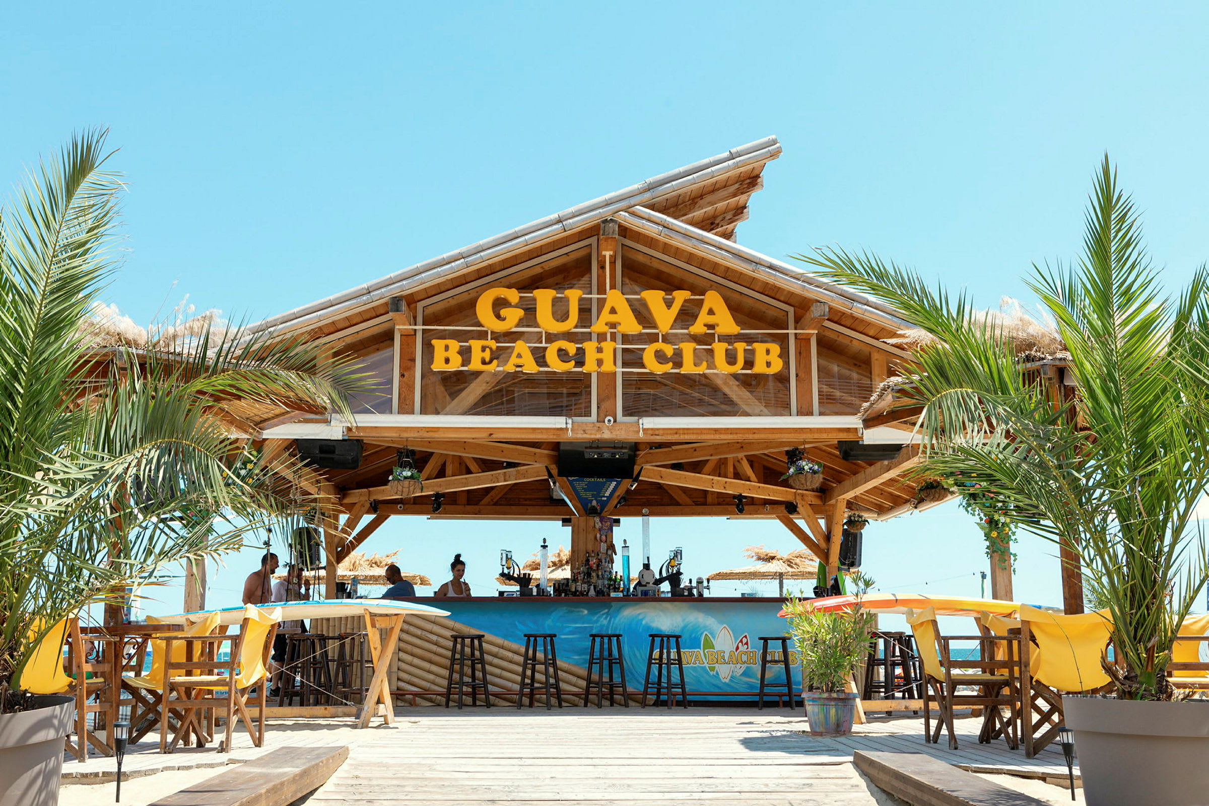 Bild av Guava Beach Club, ett träbart med sittplatser, palmträd och en klar himmel. (AI-genererad text)