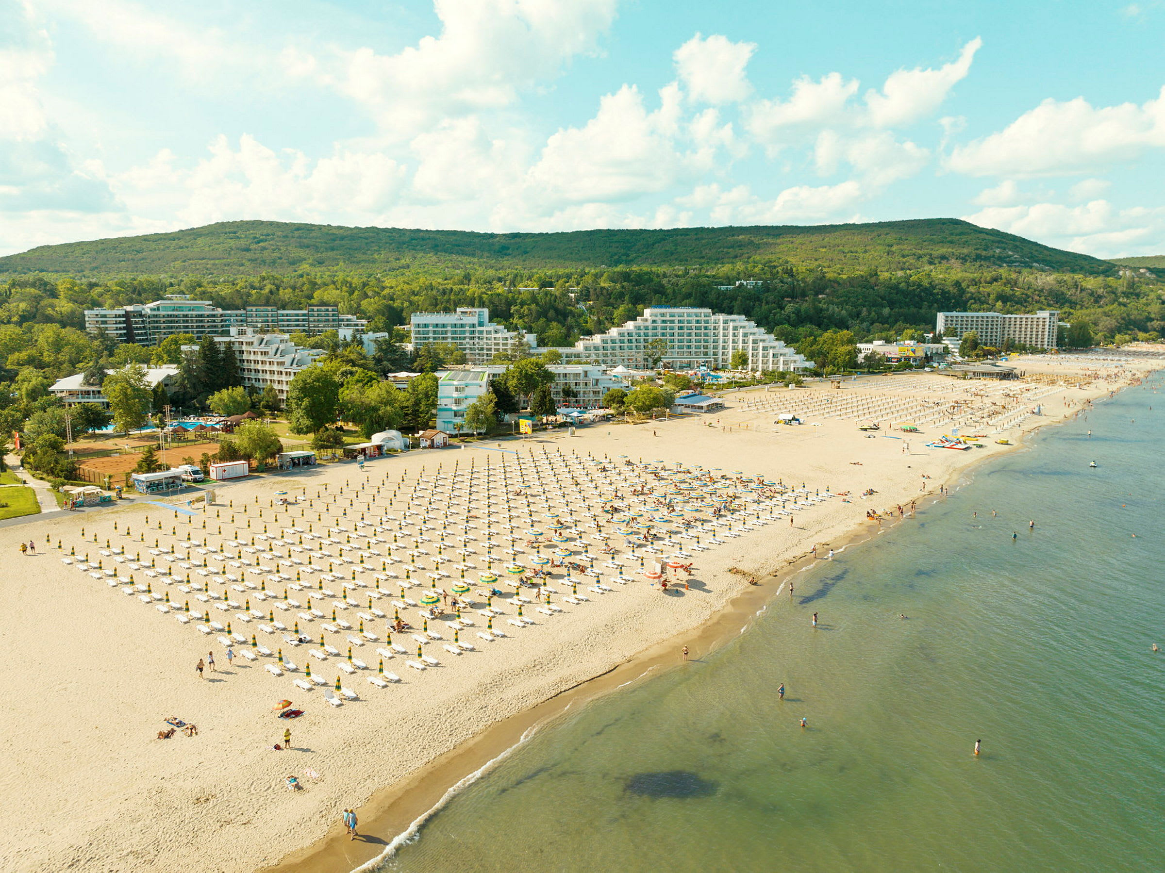 Strand med solstolar och parasoller, omgivna av hotellbyggnader och grönska. (AI-genererad text)