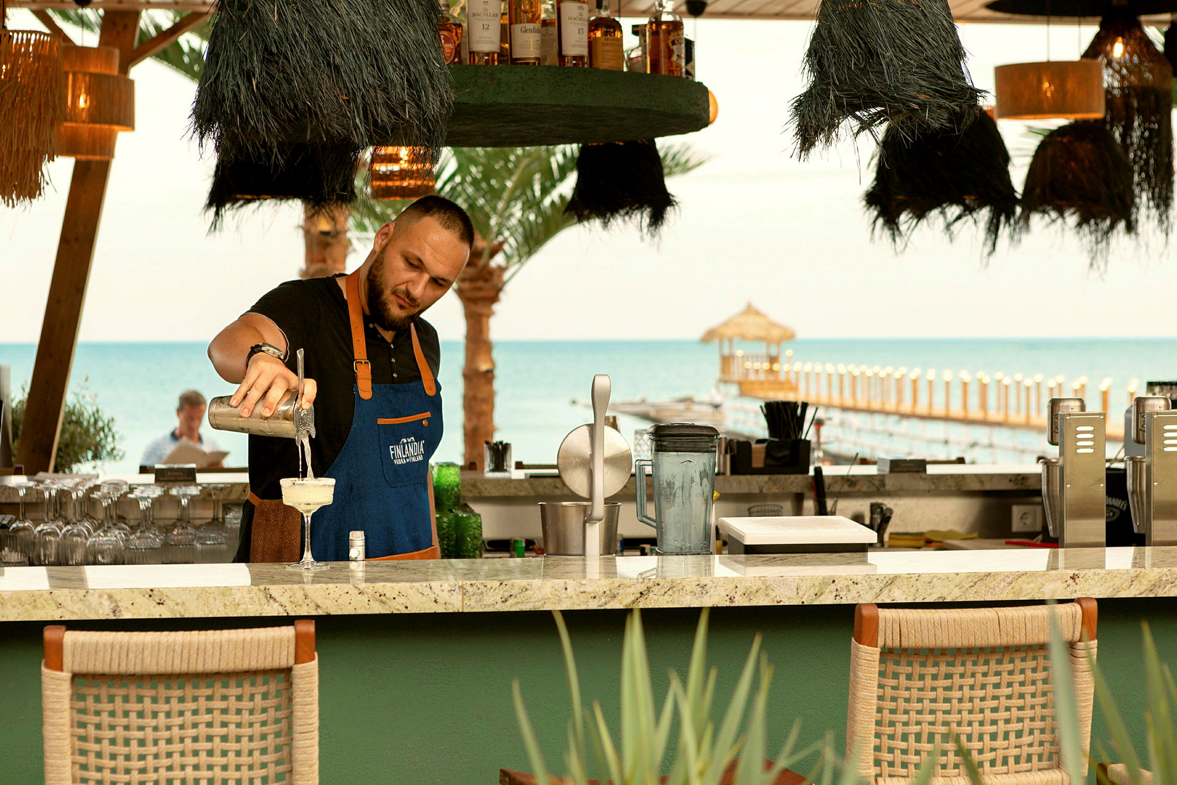 En bartender vid en strandbar häller upp en drink med havet och en brygga i bakgrunden. (AI-genererad text)
