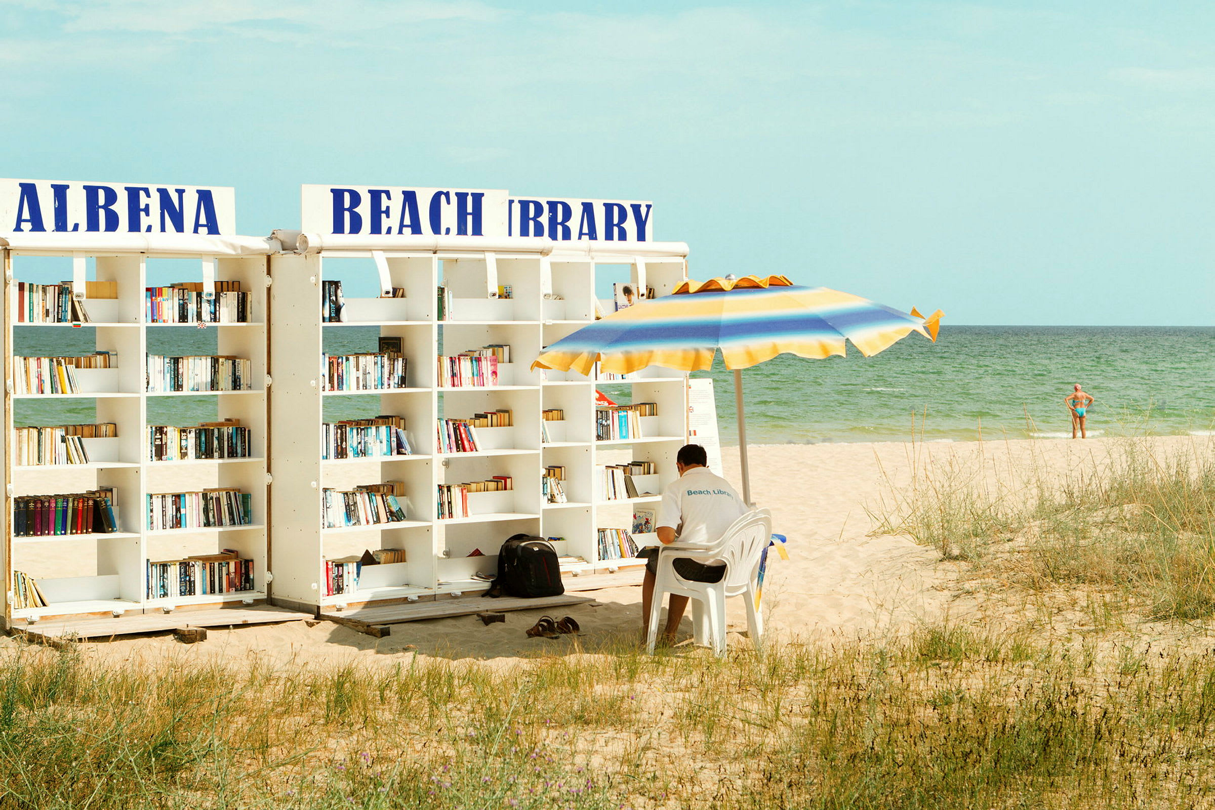 En strand med en utomhusbibliotek och en man som sitter under ett parasoll. (AI-genererad text)