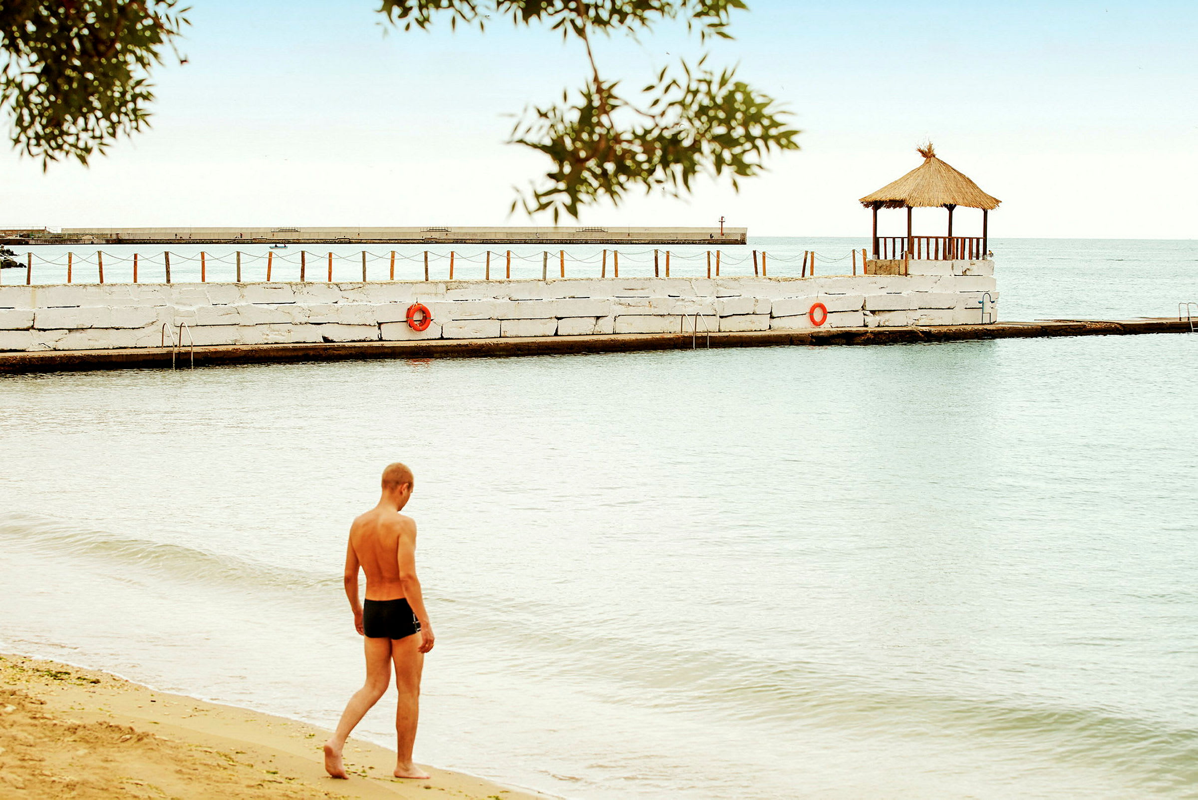 En person promenerar på en strand bredvid vatten. En paviljong står på en brygga. (AI-genererad text)