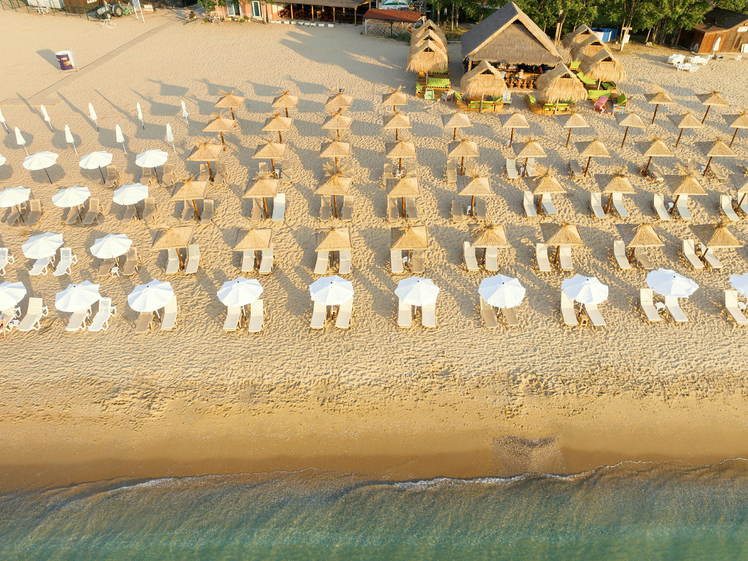 Flygfoto av en strand med radenade solstolar och parasoller, samt strandbari med halmtak i bakgrunden. (AI-genererad text)